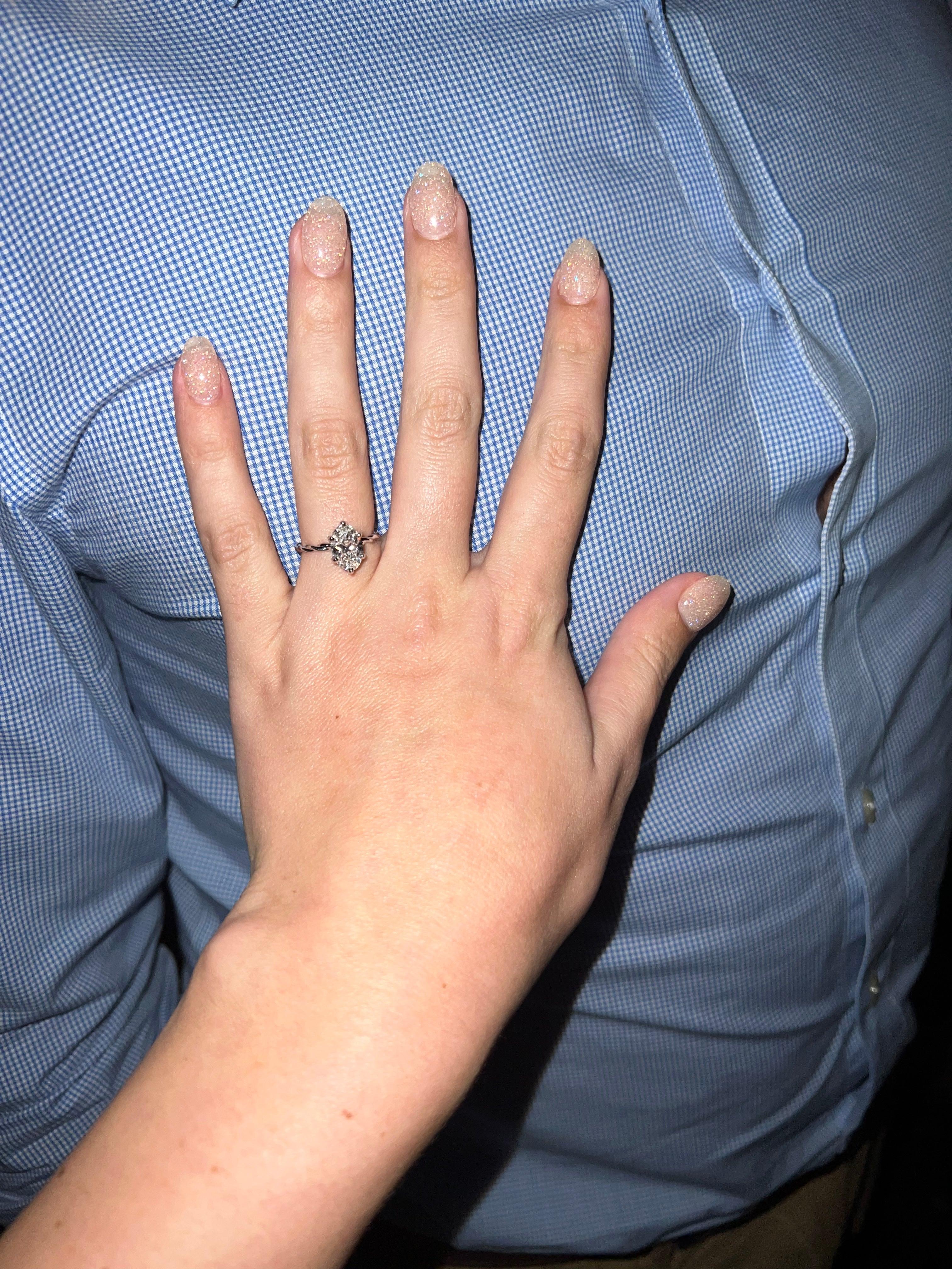 Left hand with glitter nails wearing a halo diamond engagement ring resting on a blue gingham shirt - Kennedy & Aaron
