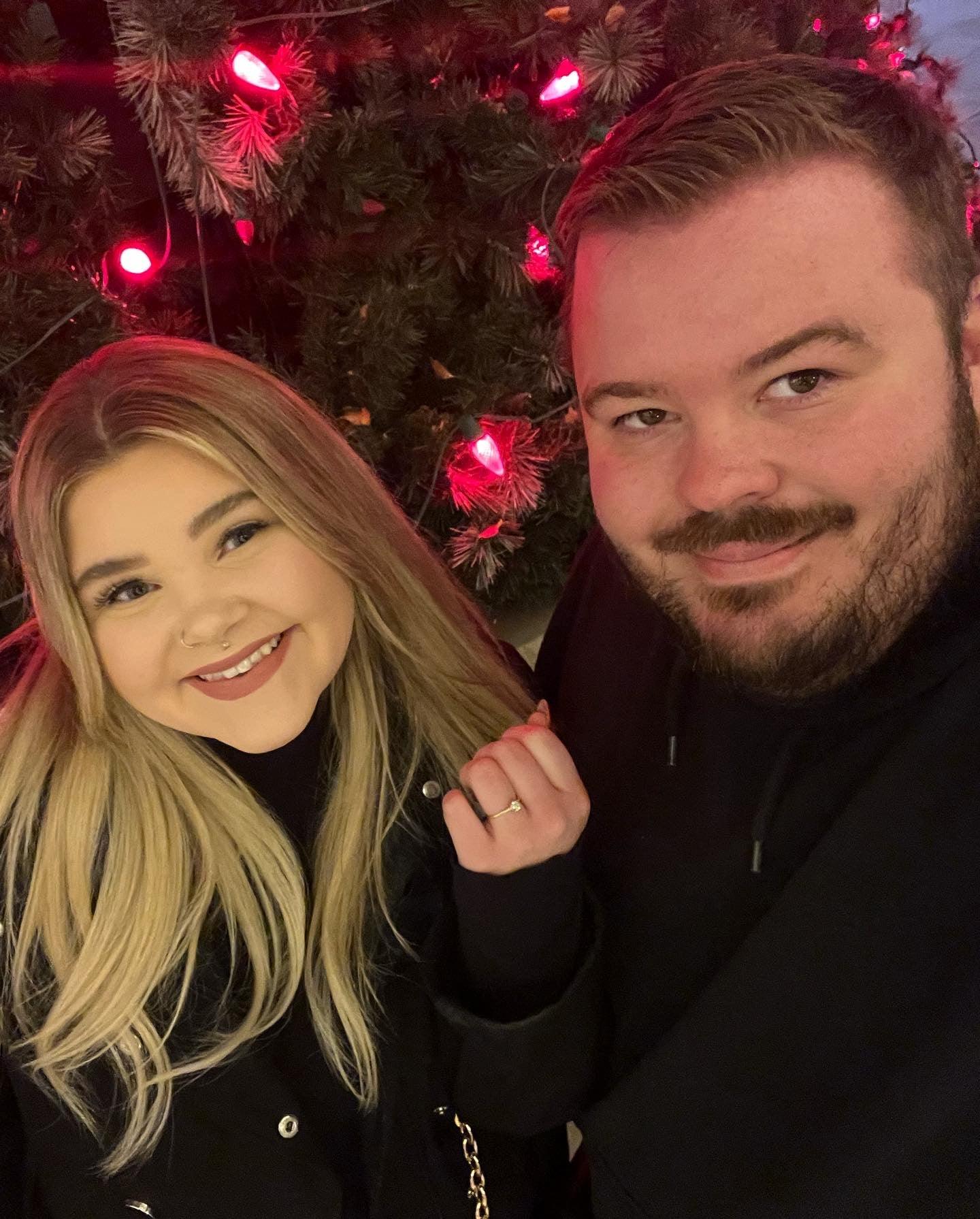 Smiling woman showing her engagement ring while posing with her partner in front of a Christmas tree with red lights