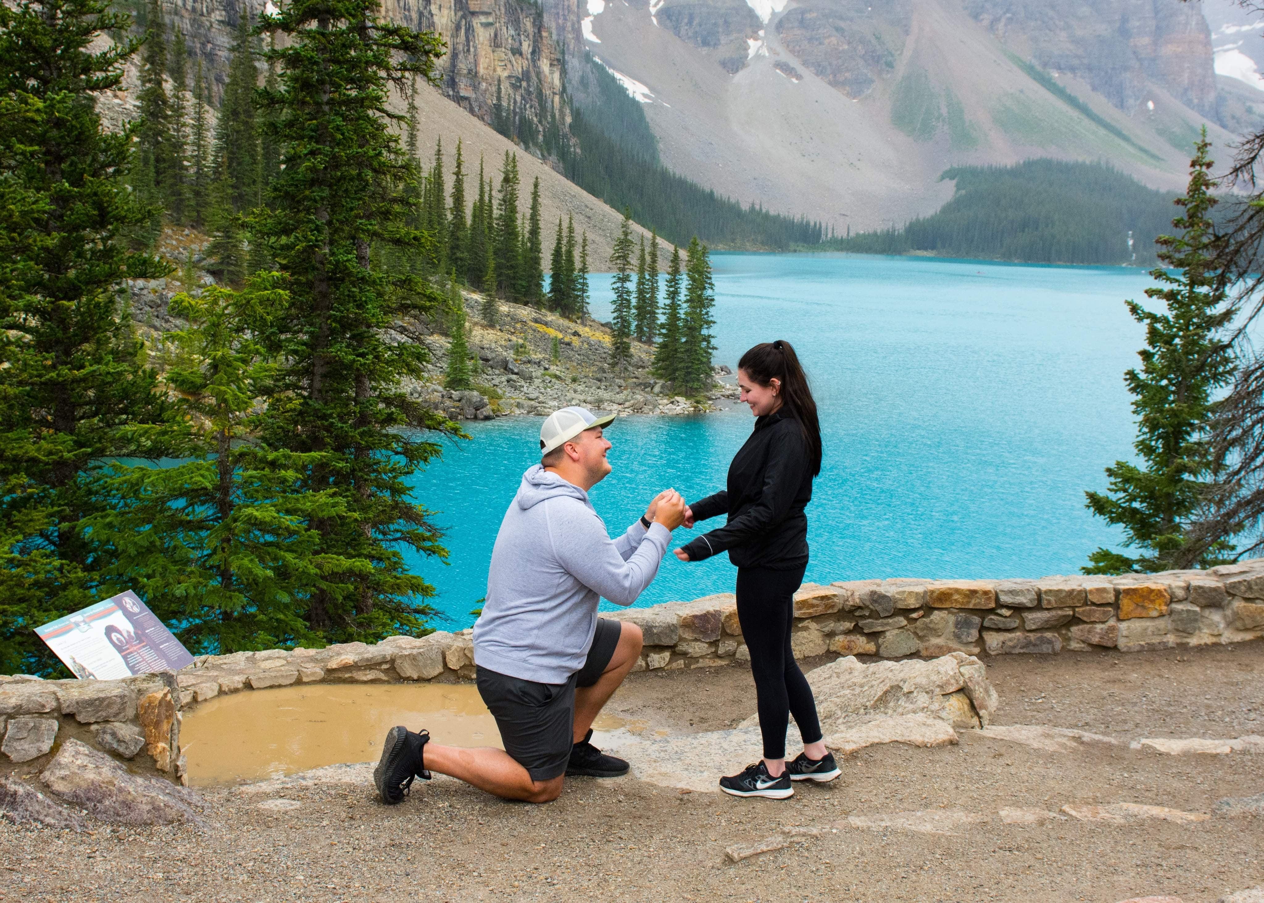 Sara and Austin: Austin kneeling, proposing to Sara beside a turquoise mountain lake and stone wall.