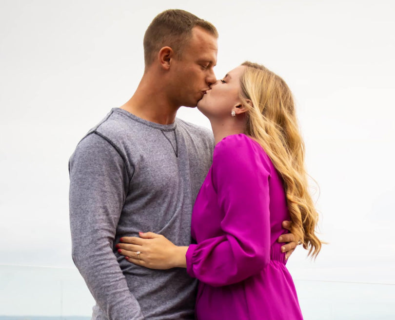 Woman and man kissing, woman in magenta dress showing engagement ring against pale sky