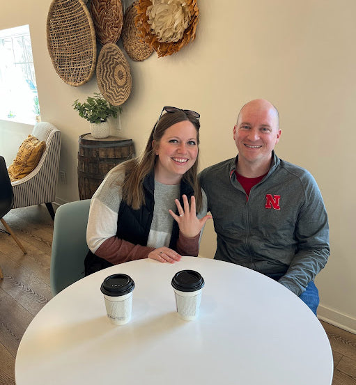 Woman showing engagement ring while seated with partner at a café table with two coffee cups and woven wall baskets in the background