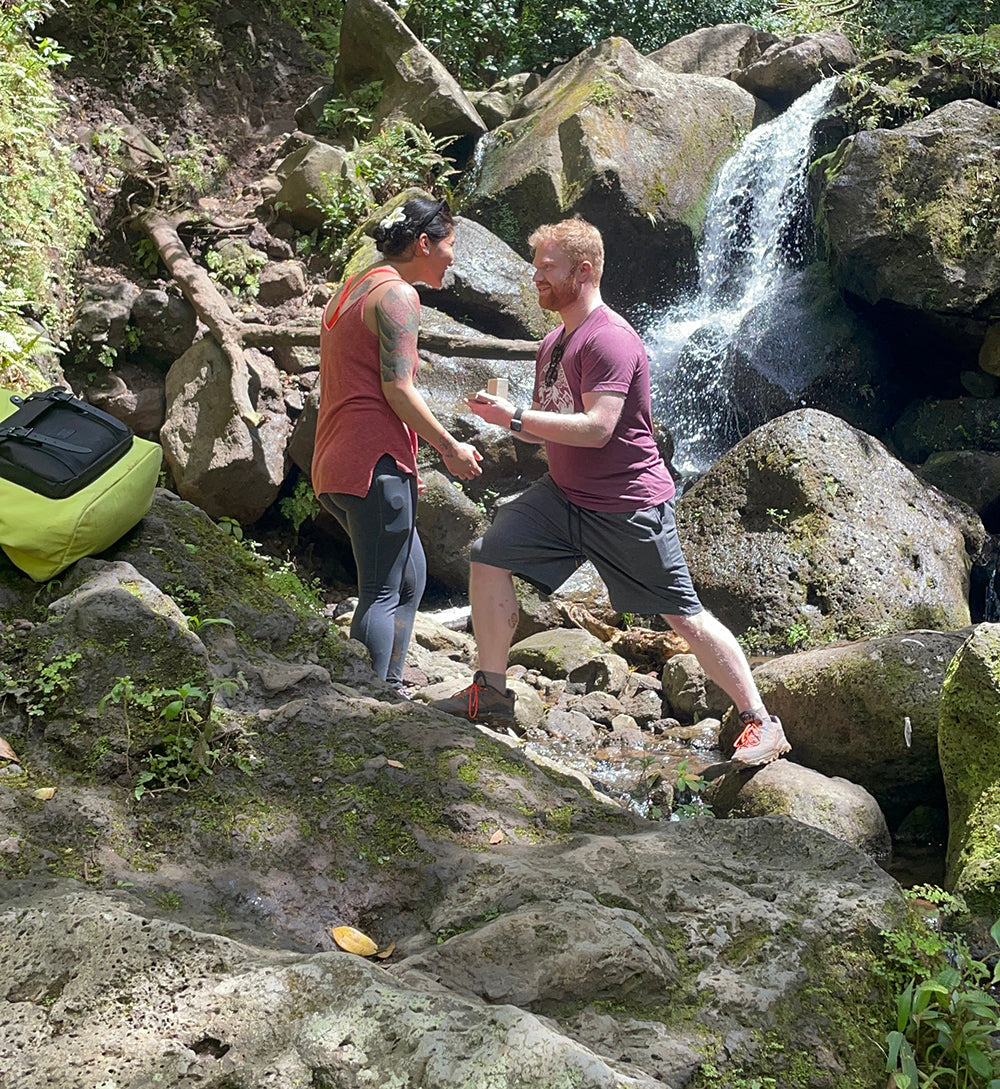 Man kneeling and proposing to partner, holding an open ring box beside a rocky waterfall