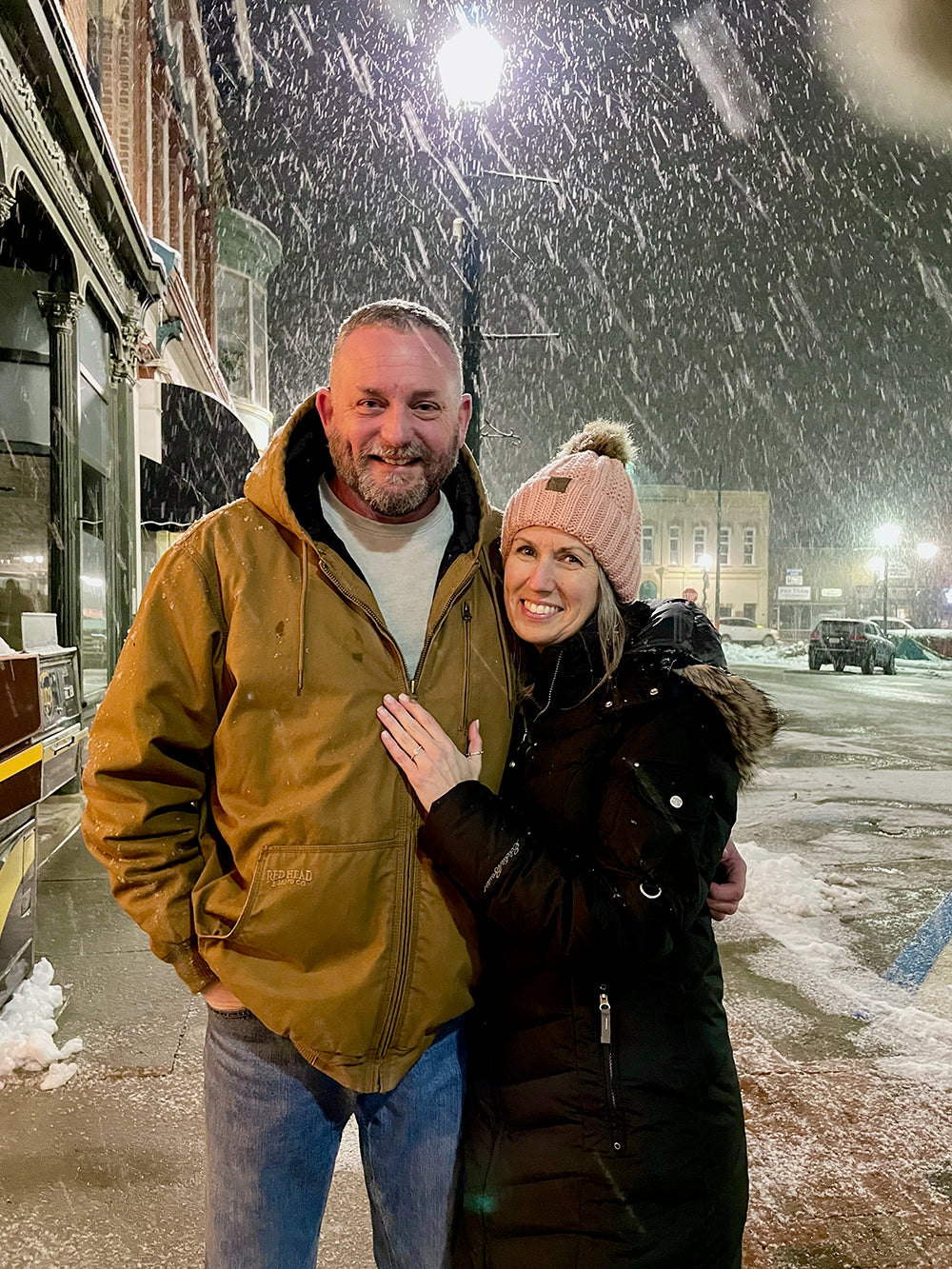 Heather and Jeff smiling in falling snow downtown, Heather shows engagement ring; man's jacket reads REDHEAD.
