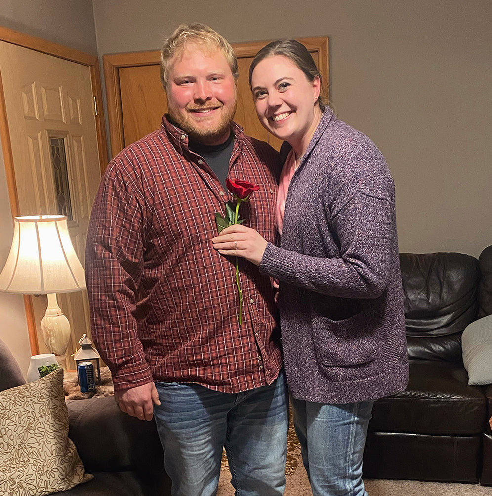 Engaged couple smiling together, woman holding a single red rose in a living room with lamp and couch