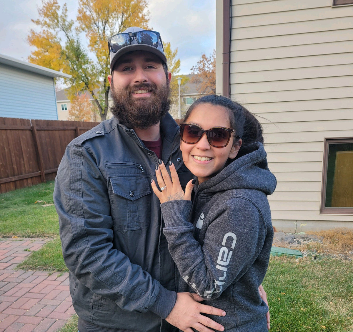 Woman showing her engagement ring while hugging her partner in a backyard with fall trees