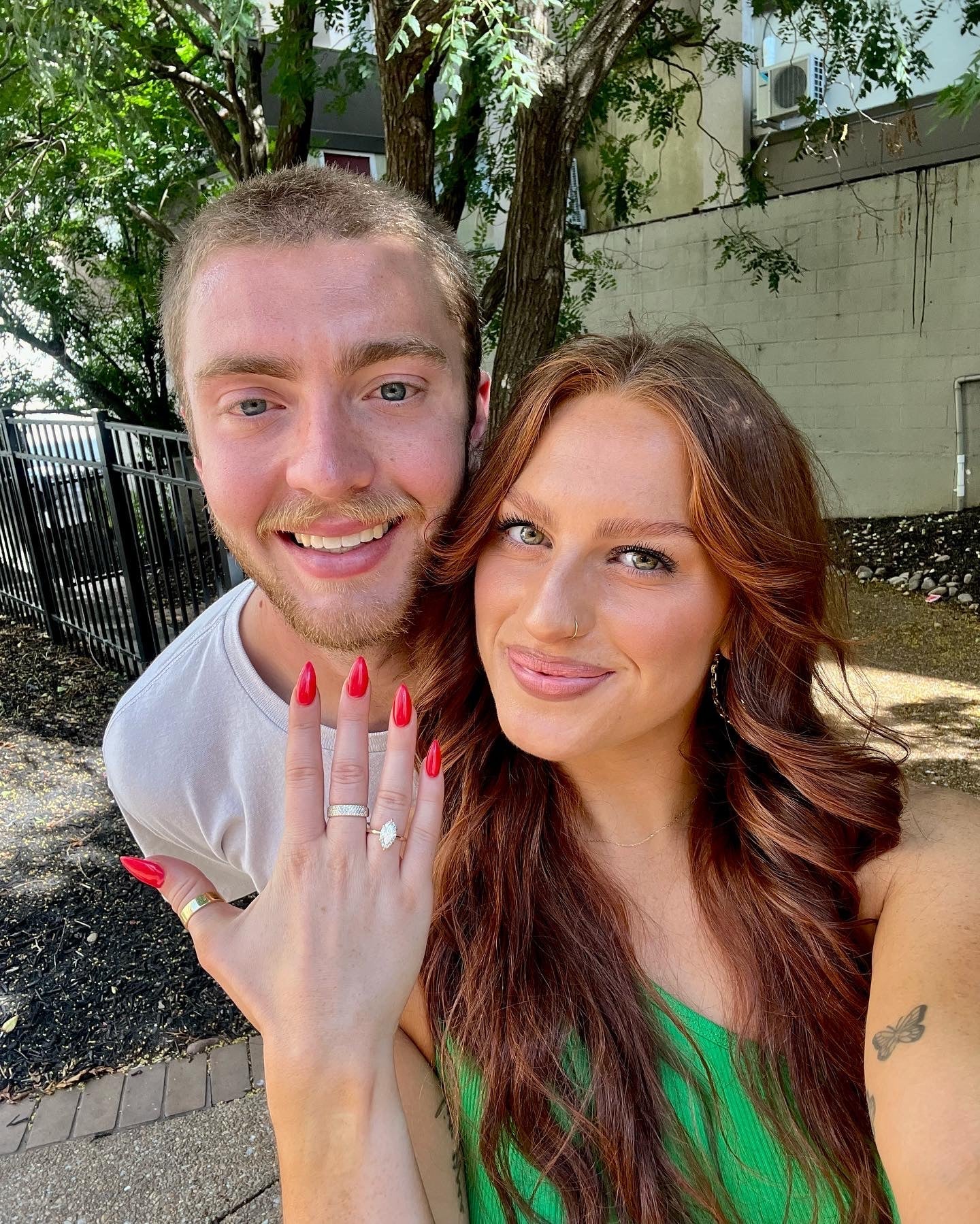 Kelsey and Will smiling in an outdoor selfie; Kelsey displays her engagement ring and red nails.