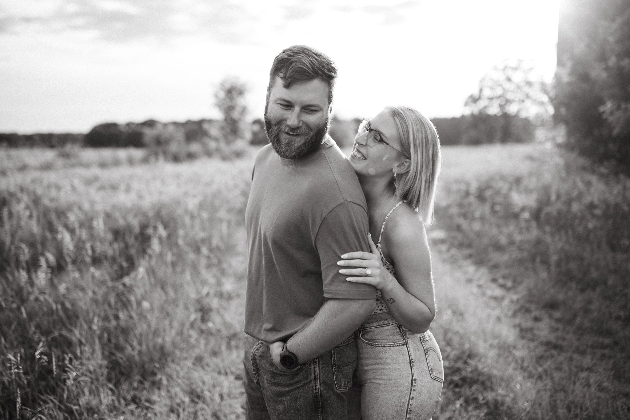 Couple embracing on a grassy path at sunset, woman smiling and showing her engagement ring