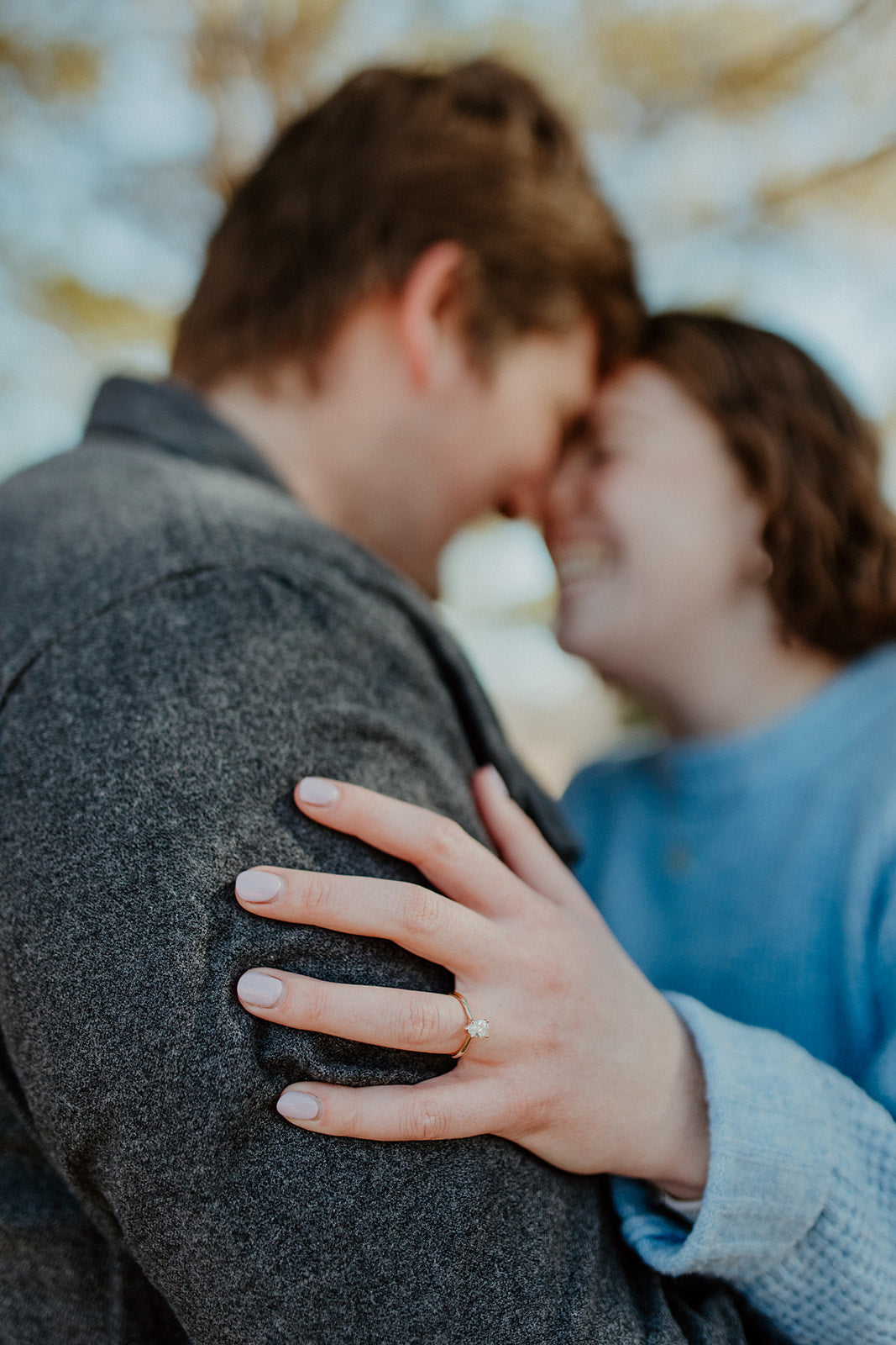 Couple smiling nose to nose as woman rests hand with pear cut yellow gold diamond ring on partner’s shoulder outdoors