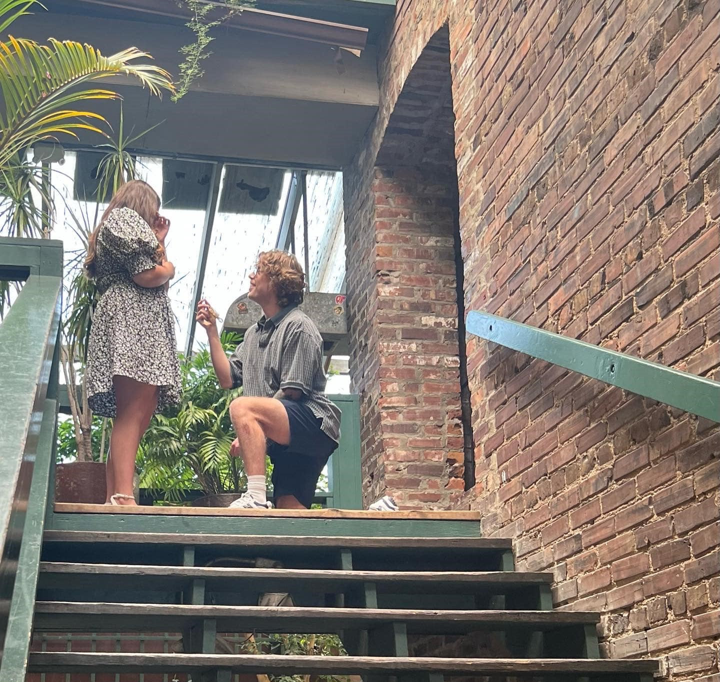 Man kneeling and offering engagement ring to woman on indoor stairwell with exposed brick wall