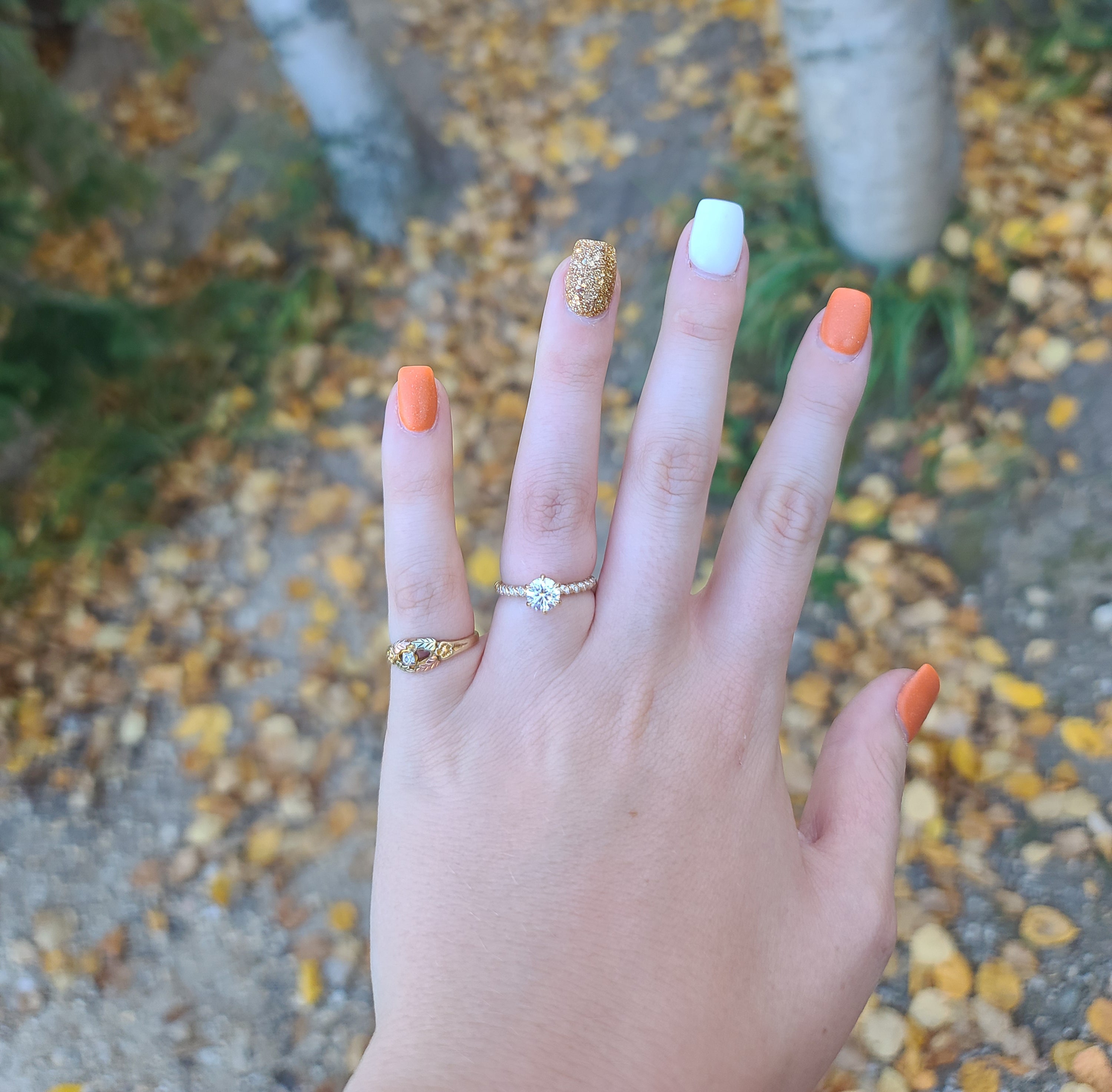 Left hand showing solitaire engagement ring and two gold rings, orange and gold glitter nails over fallen autumn leaves