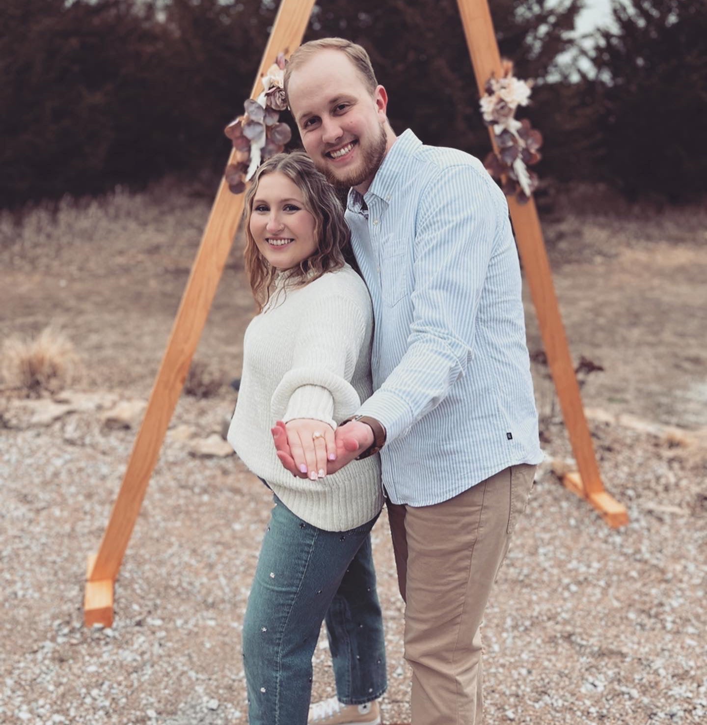 Tori and Jackson standing under a wooden arch, Tori showing an engagement ring on her outstretched hand