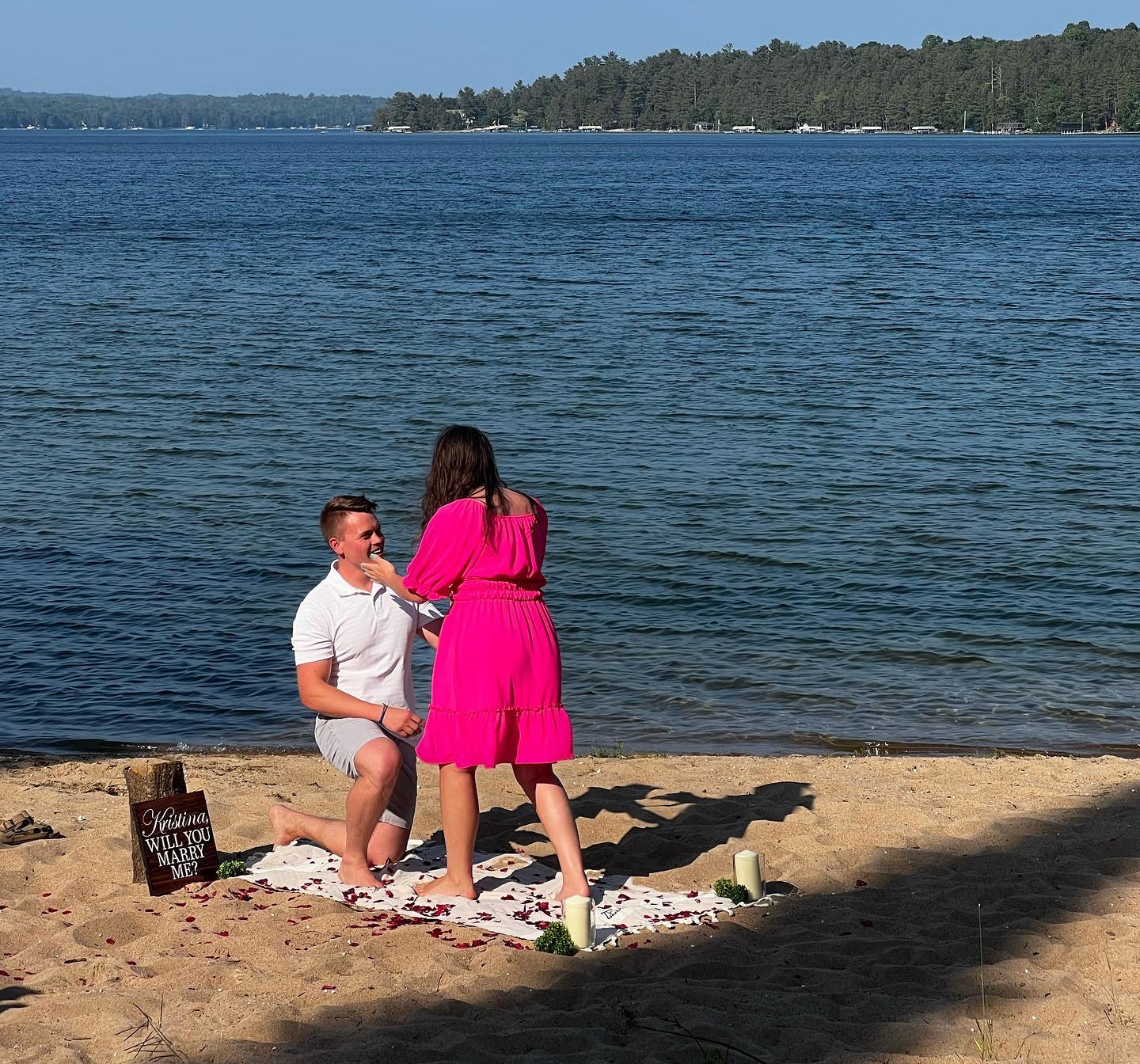 Man kneeling to propose to Kristina on a lakeside beach; wooden sign reads 'Kristina WILL YOU MARRY ME?'