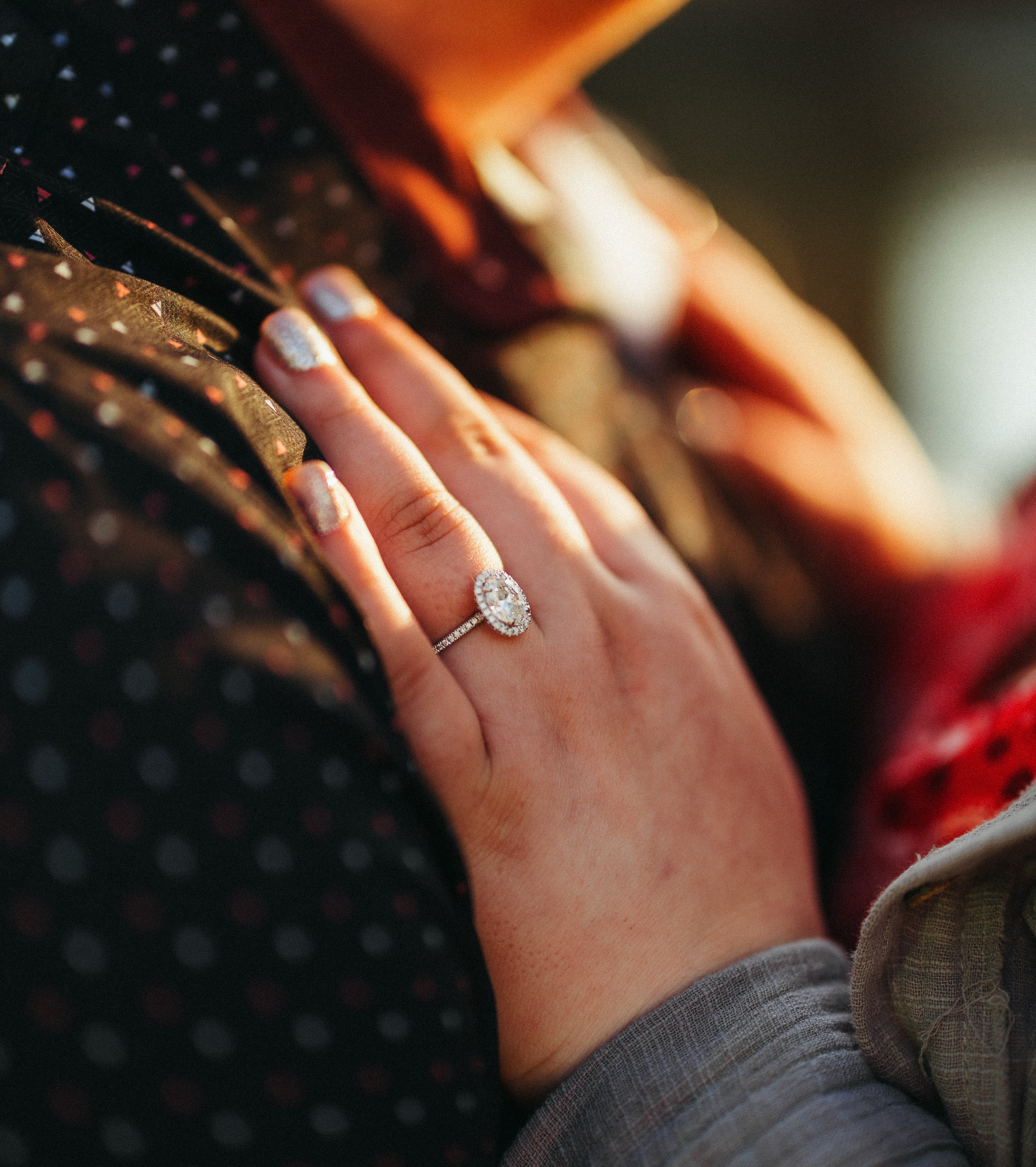 Morgan's hand wearing an oval halo engagement ring resting on Cody's shoulder in warm sunlight