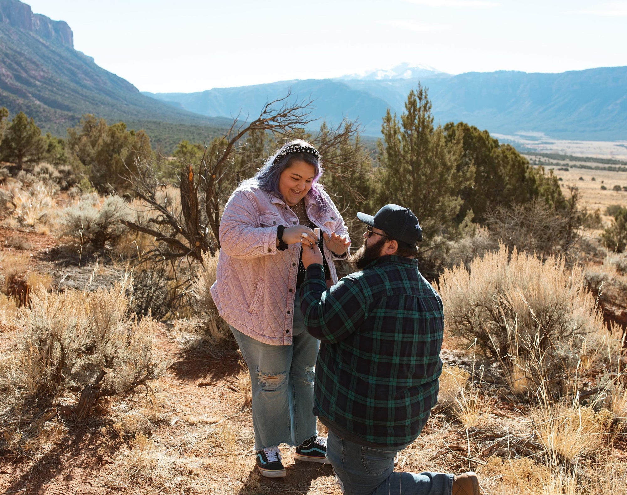 One person kneeling to propose to their partner amid sagebrush and red-rock desert mountains
