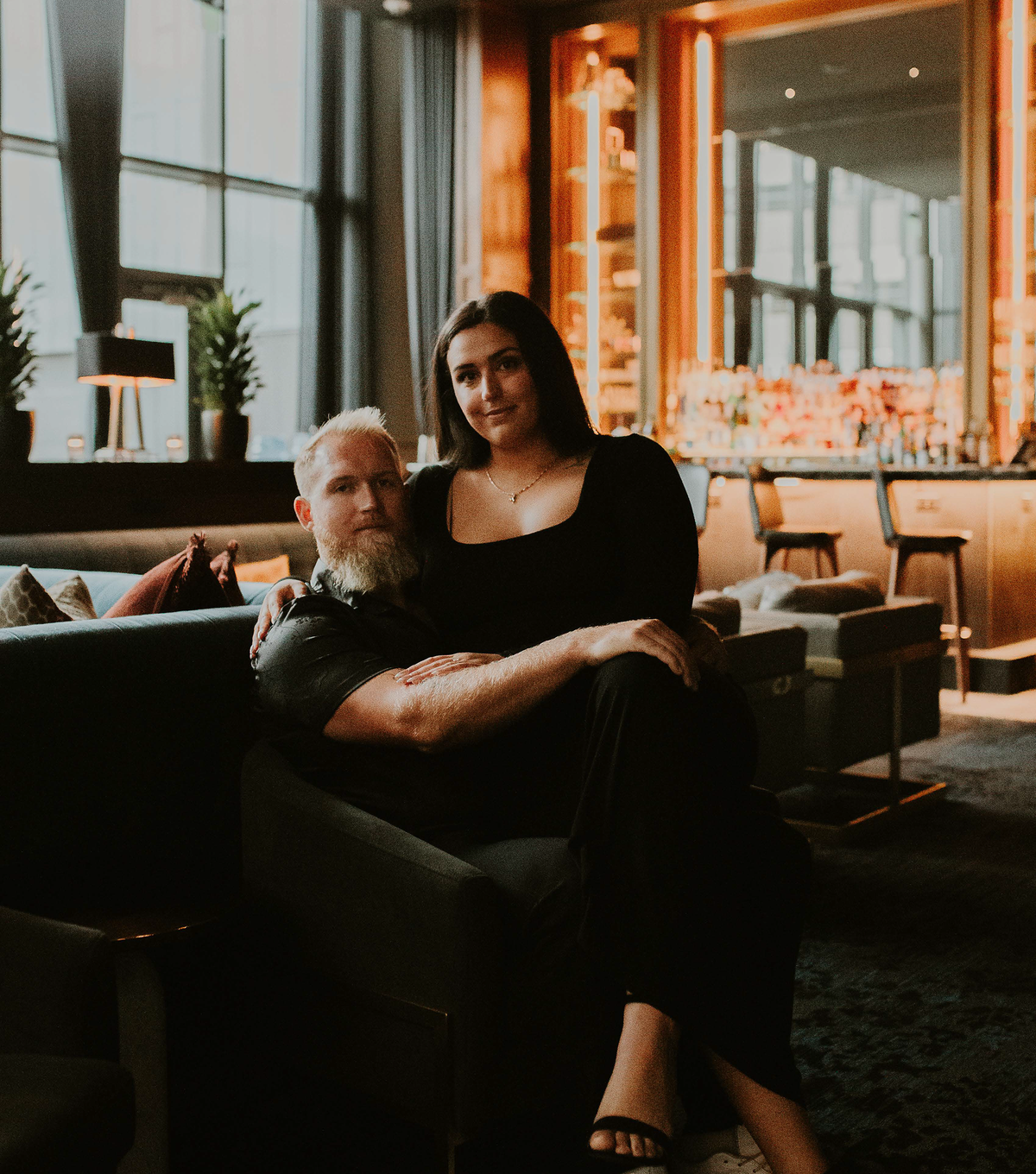 Man seated on a sofa with woman standing behind him, posing in a hotel lounge with warm bar lighting
