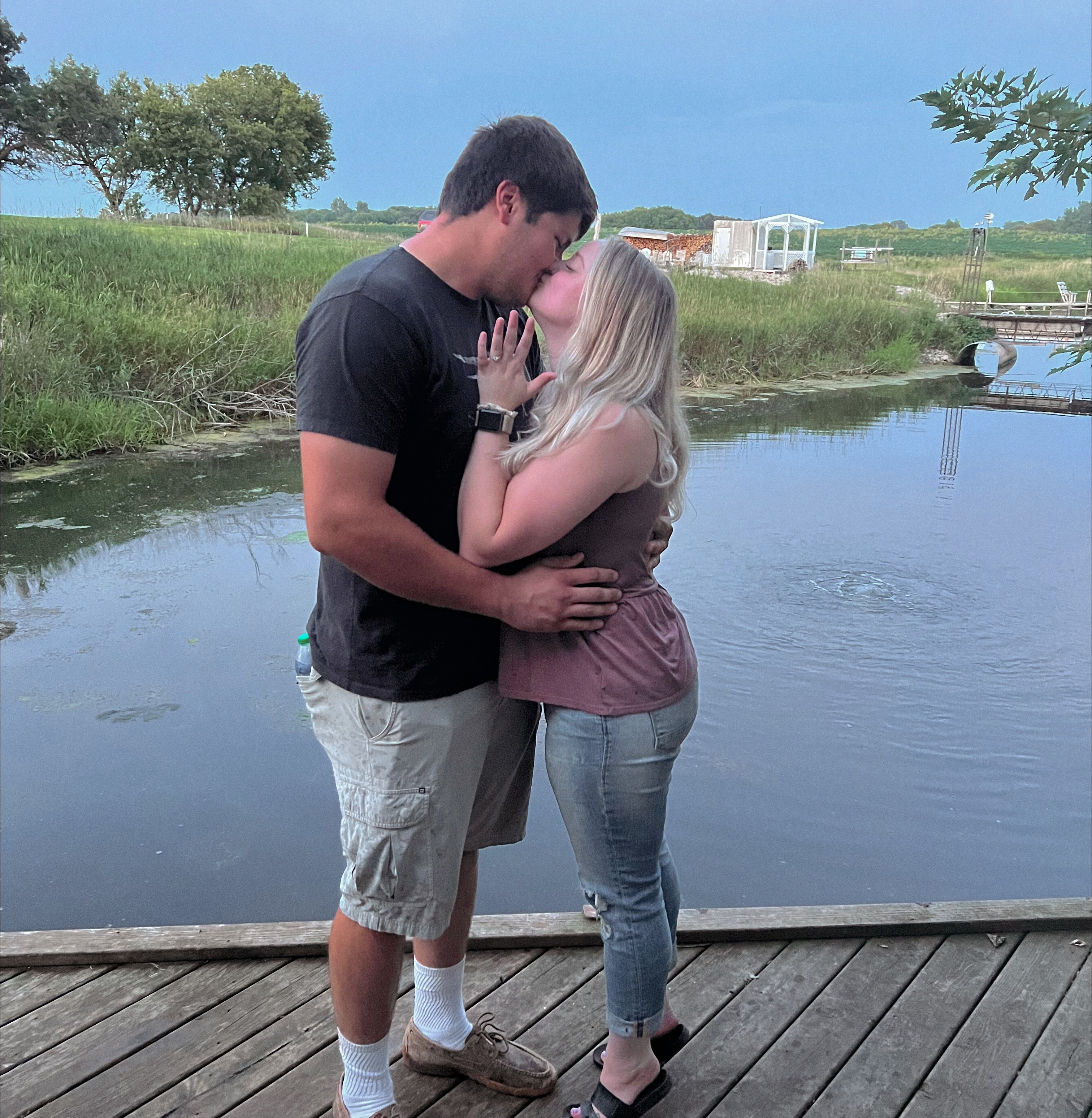 Couple kissing on a wooden dock by a pond at sunset, woman showing her engagement ring