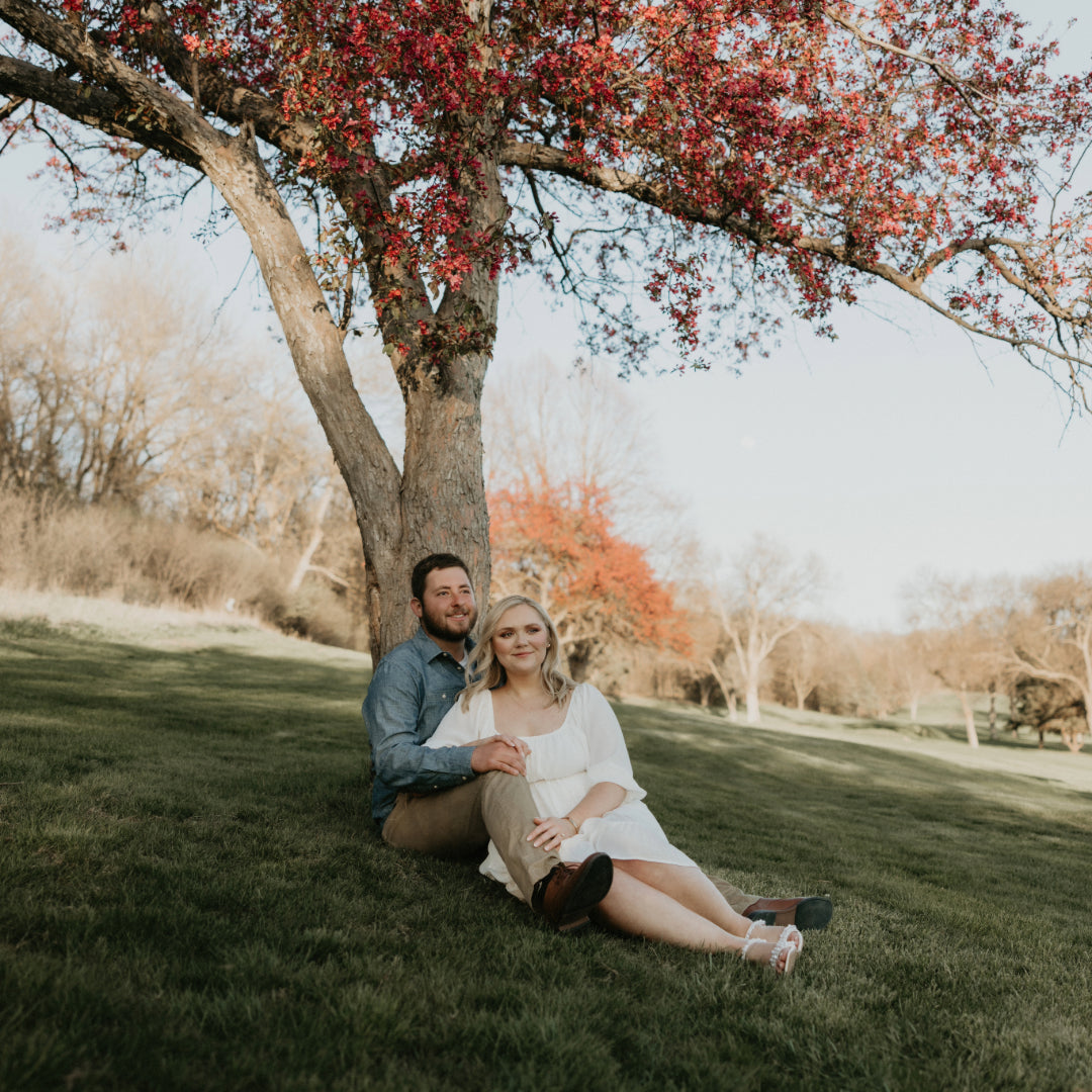 Kelsey and Christopher seated on grass, leaning against a tree with red blossoms