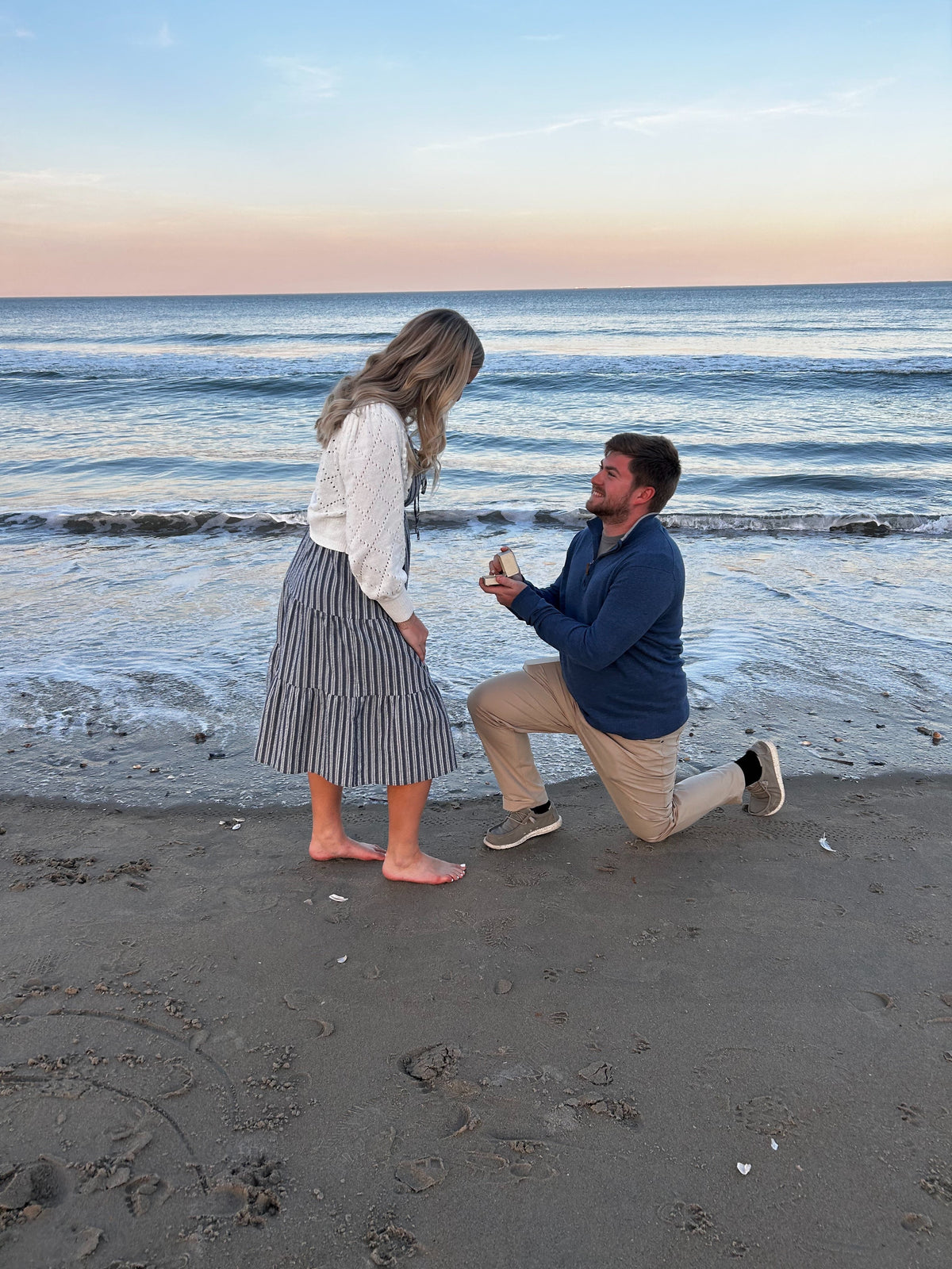 Man kneeling on a beach at sunset offering an open box with an oval diamond engagement ring to a woman