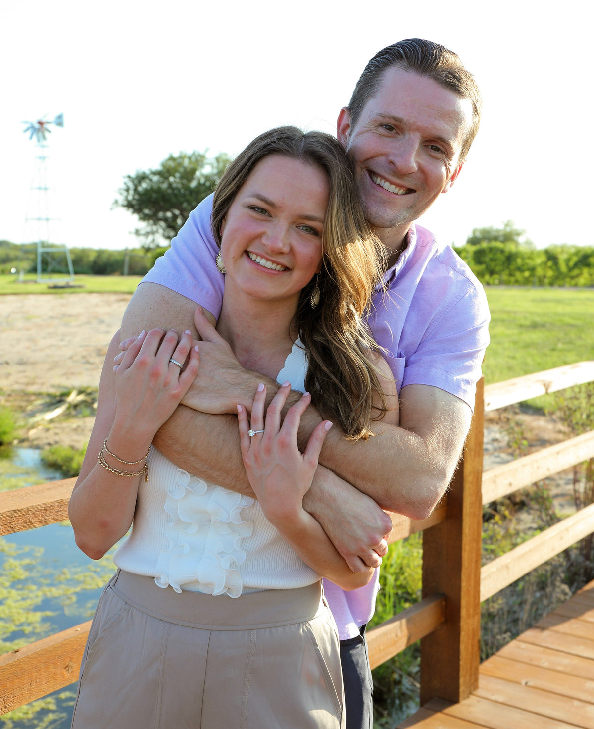 Couple embracing on a wooden bridge, woman showing 2.01 carat solitaire engagement ring with windmill and fields in the background