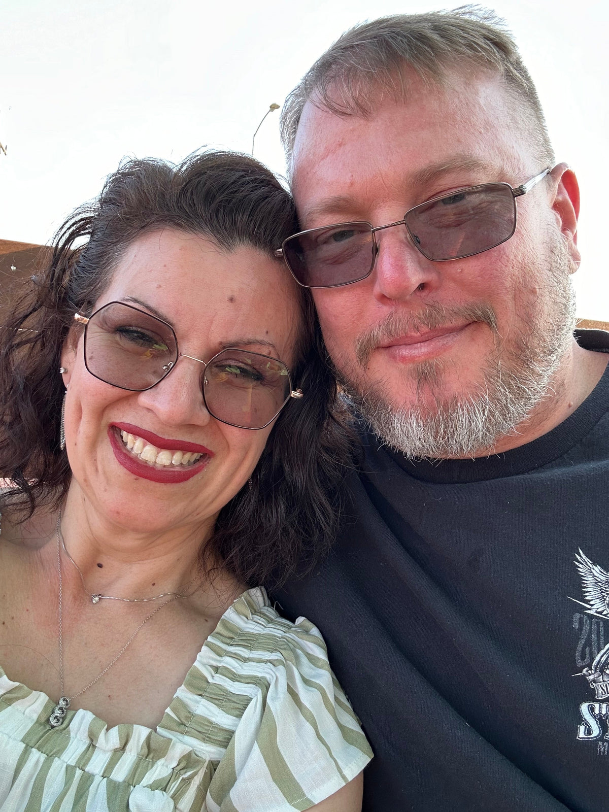 Couple smiling in a close-up selfie wearing sunglasses and leaning together outdoors