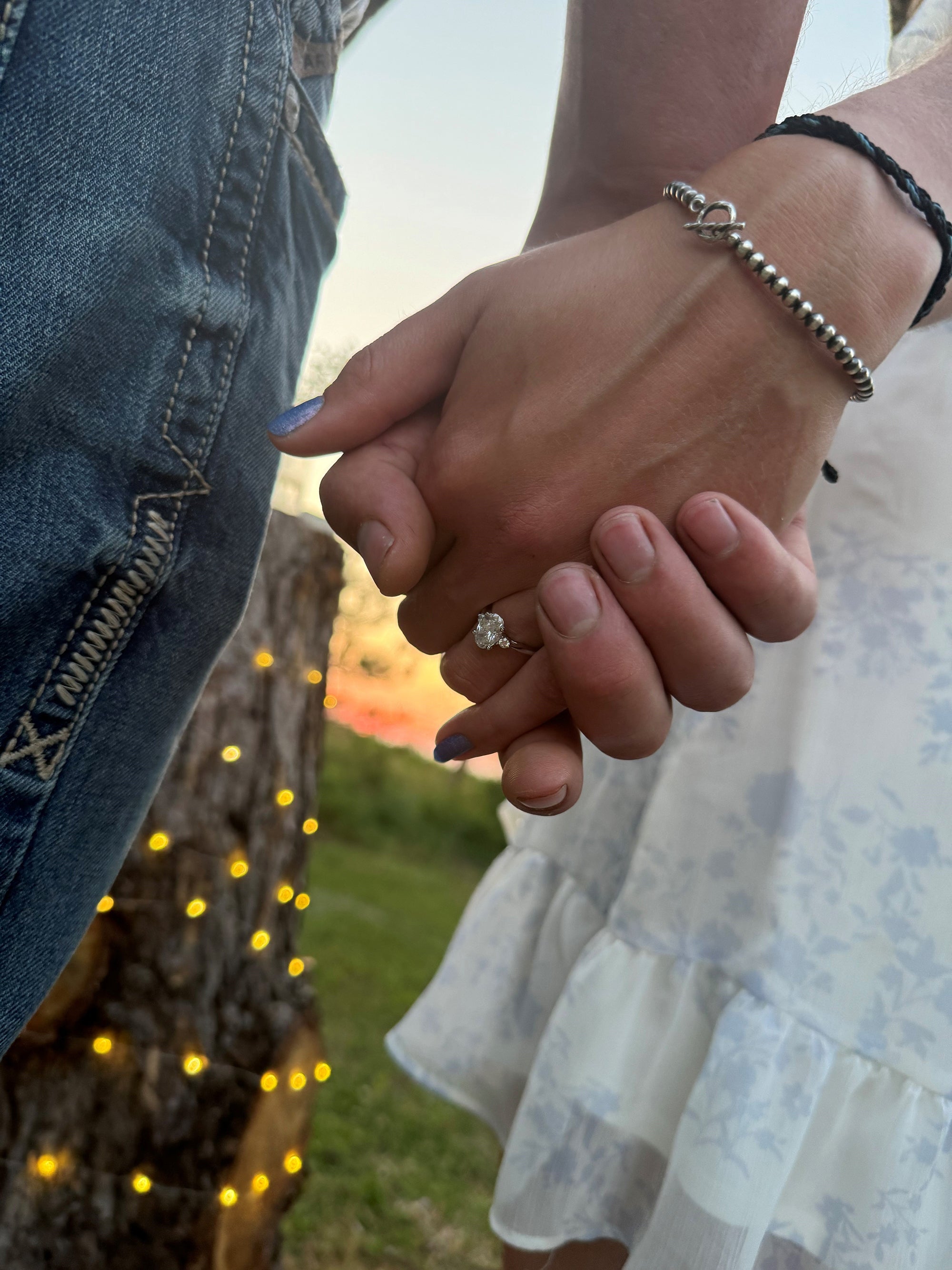 Jen and Dane holding hands, engagement ring on woman's finger, sunset background with string lights on tree