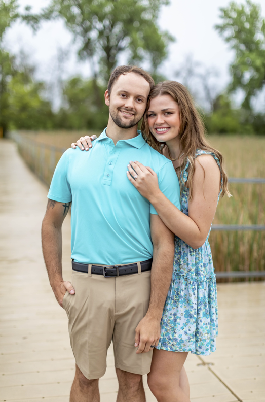 Smiling couple on a wooden boardwalk, woman resting her hand with an engagement ring on the man’s chest, tall grasses and trees behind them