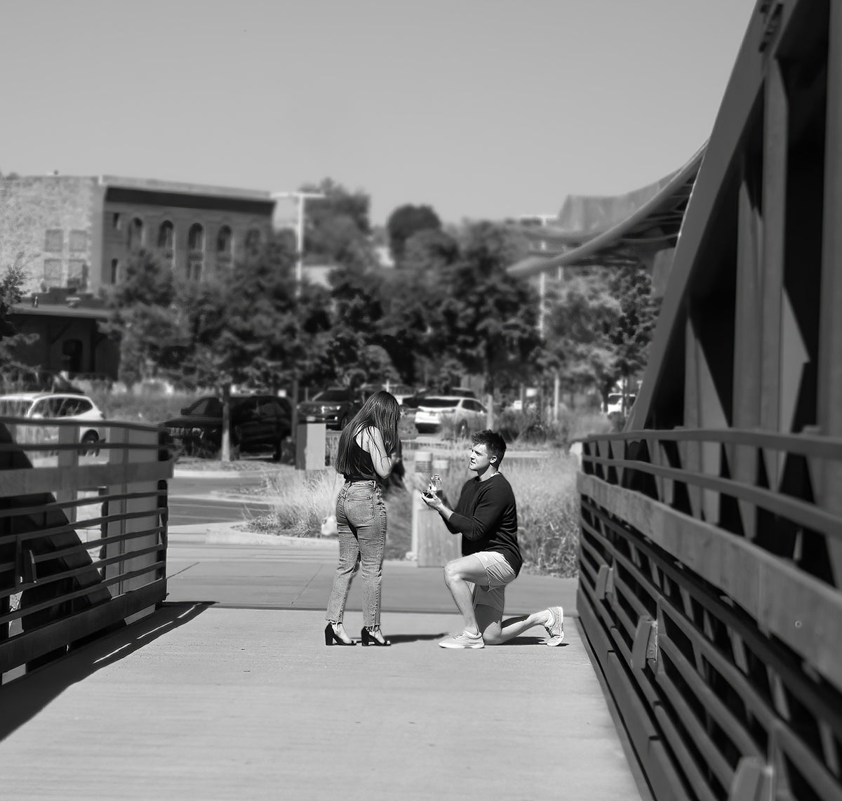 Man kneeling and presenting a ring to a woman on a pedestrian bridge during a daytime proposal.