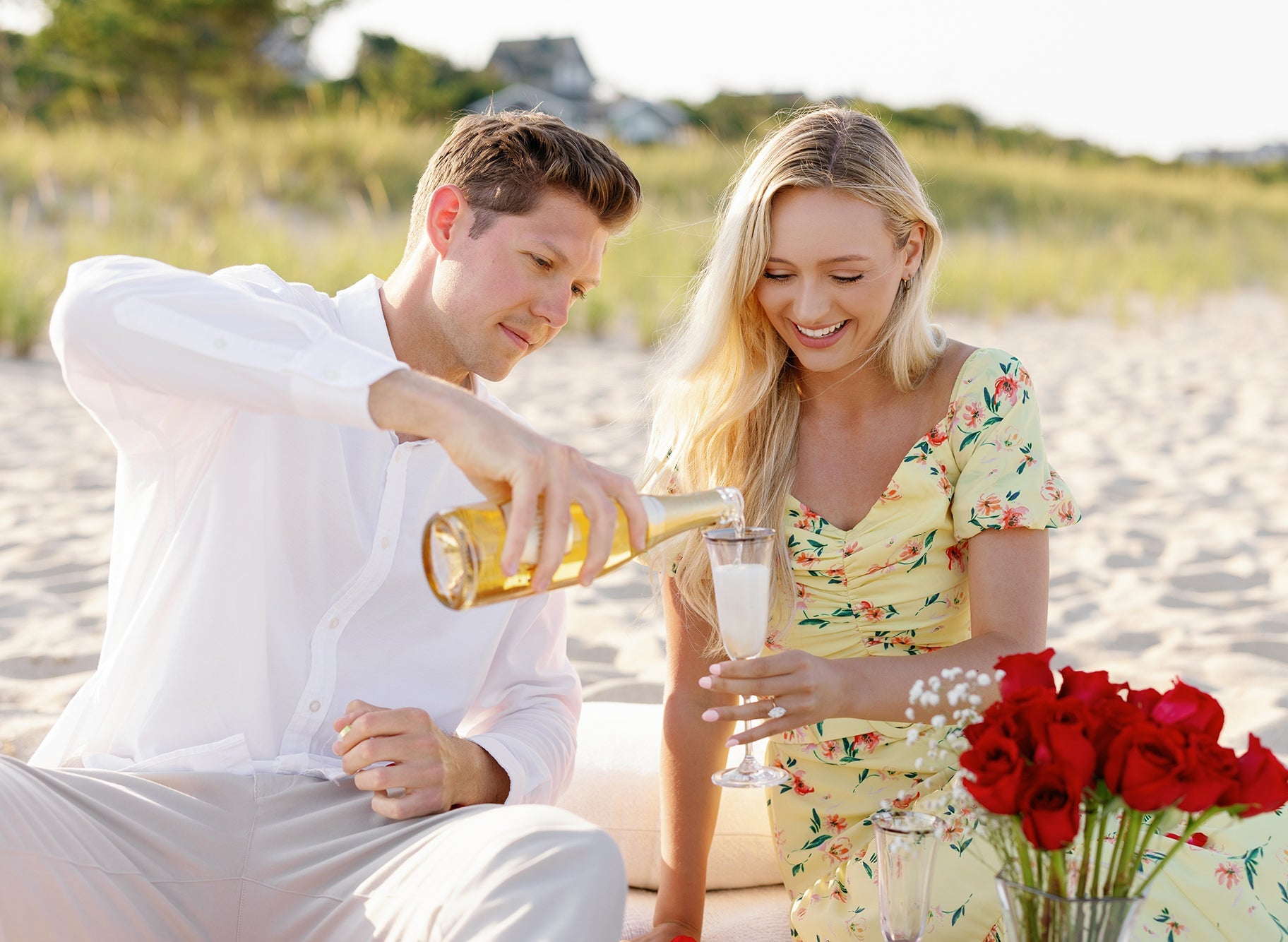 Erica and Morgen at a beach picnic; he pours champagne as she admires her engagement ring