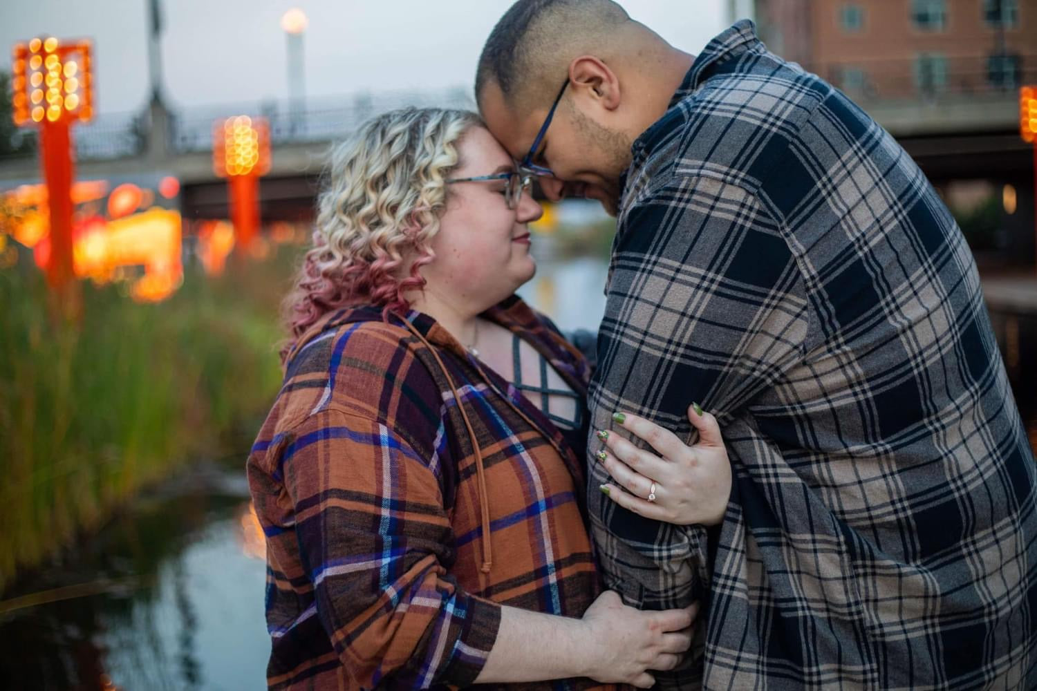 Couple touching foreheads in an embrace by a downtown Omaha park waterway at dusk, woman in brown plaid showing engagement ring