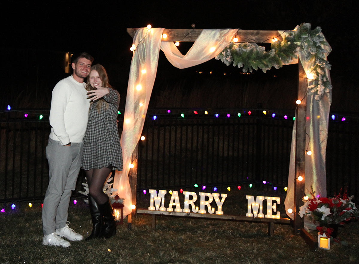 Callie and Cameron under a lighted arch with marquee letters reading MARRY ME, Callie showing engagement ring.