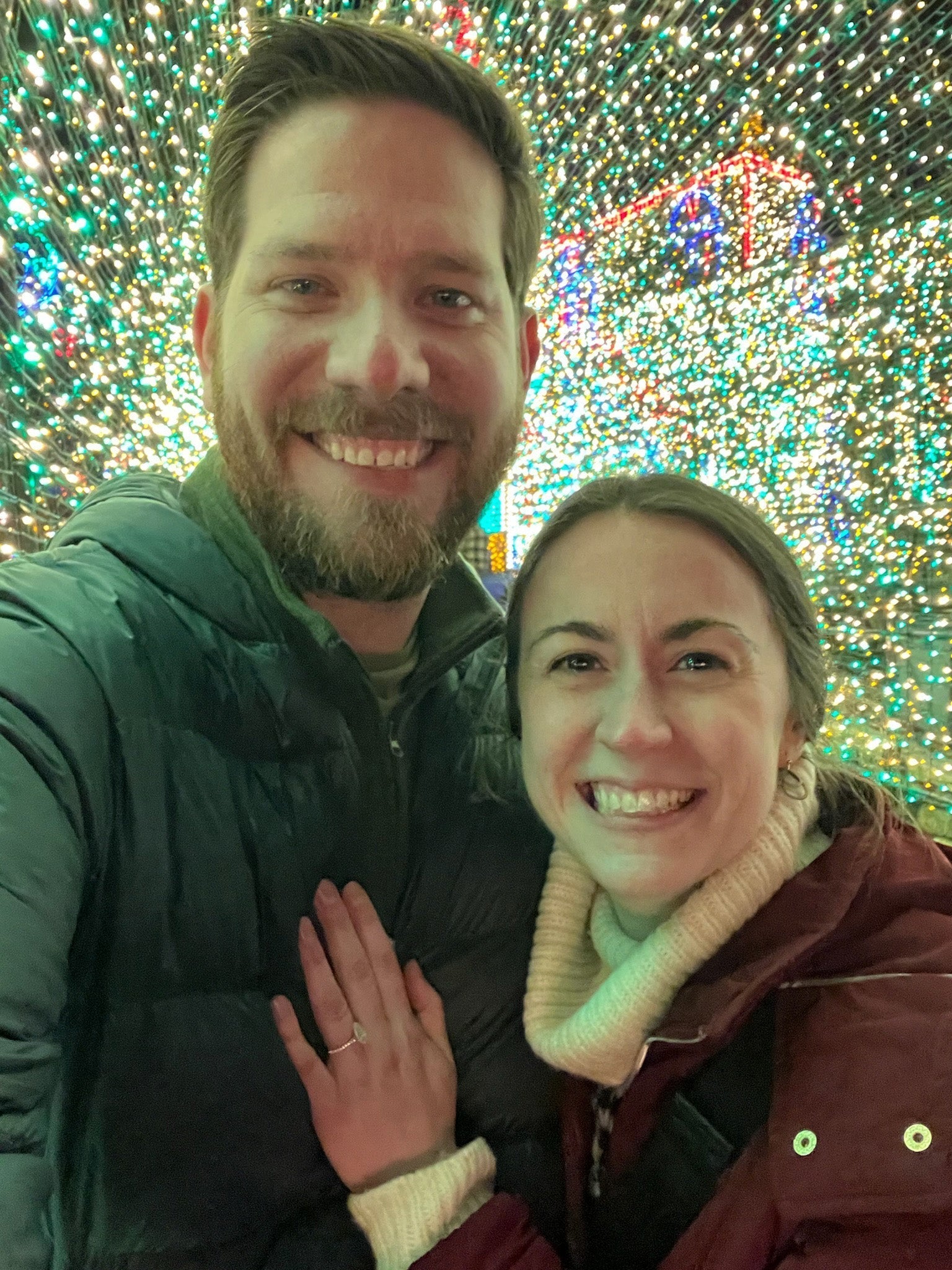 Christine smiling with hand showing engagement ring on Jake's chest, both in front of dense holiday string lights