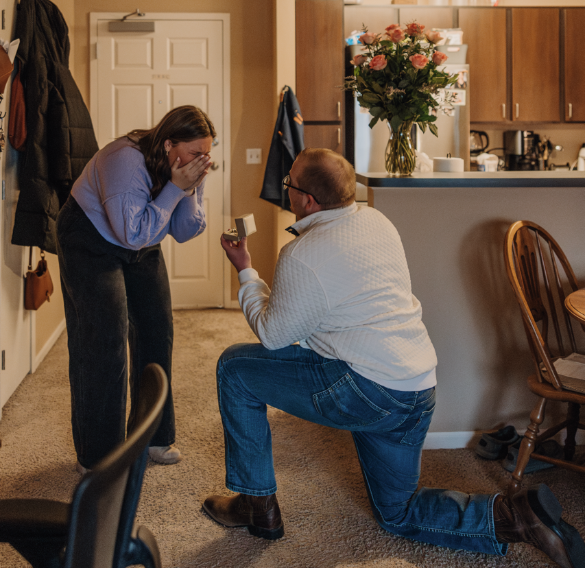 Man kneeling and proposing to his partner in a living room, she covers her face in surprise