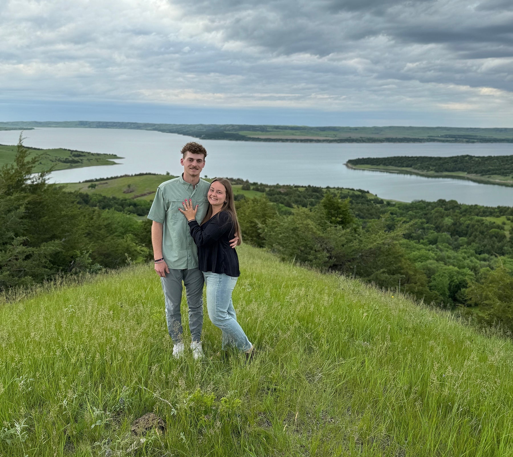 Libby and Maxwell standing on a grassy hill above a lake, Libby showing her engagement ring
