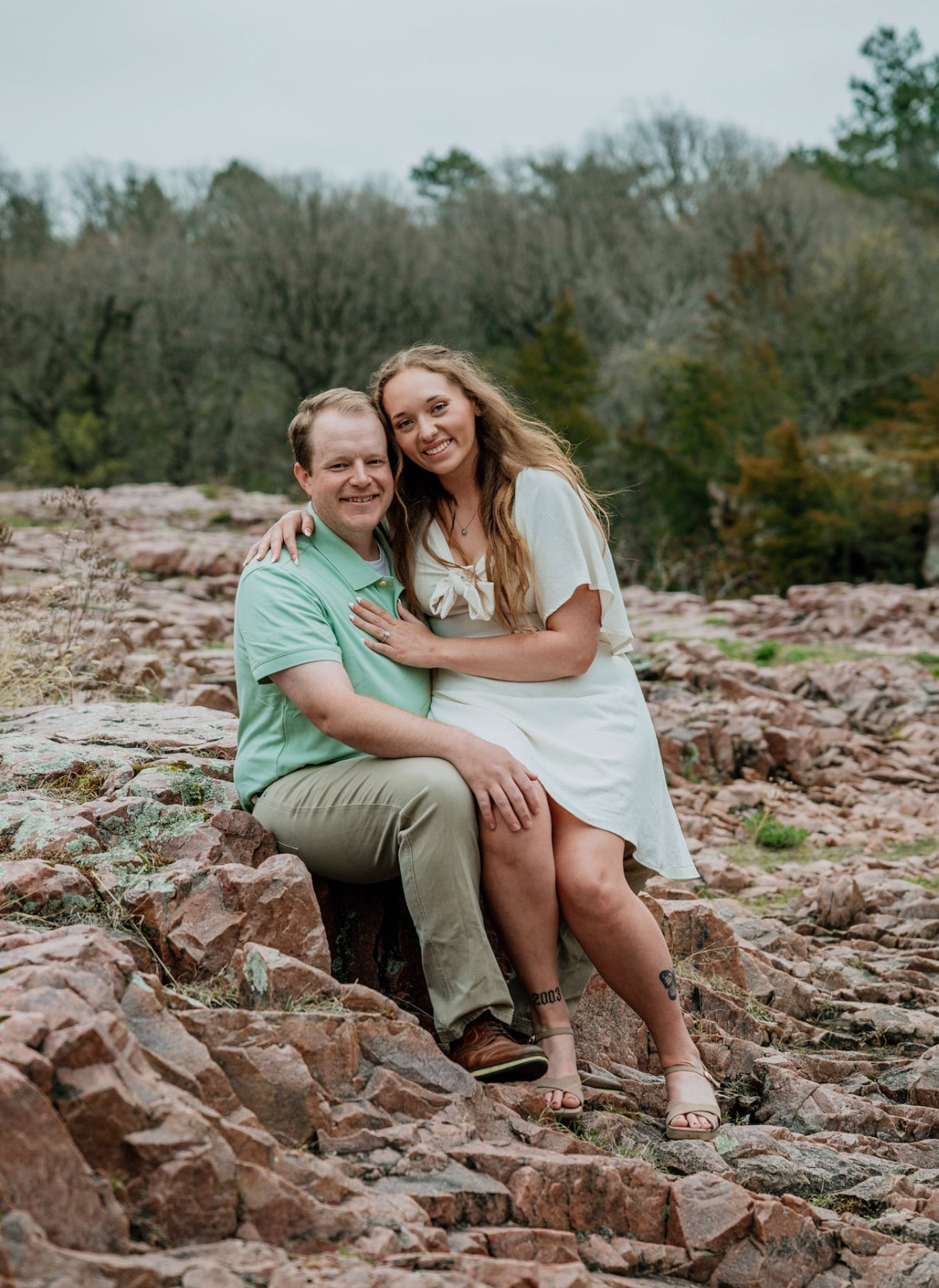 Kayley and Andrew seated on a rocky riverbank; Kayley leans on Andrew, showing her engagement ring; ankle tattoo reads 2003