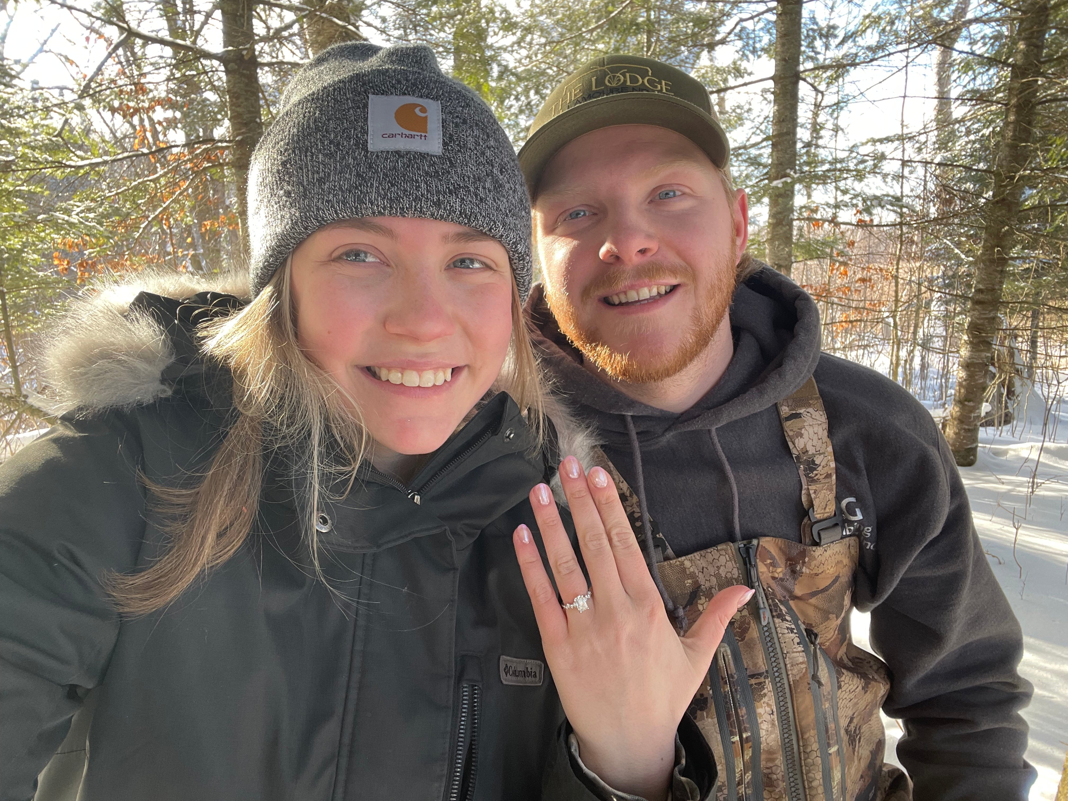 Smiling couple showing engagement ring, woman in gray knit hat and black parka, man in camouflage bibs in a sunny snowy forest