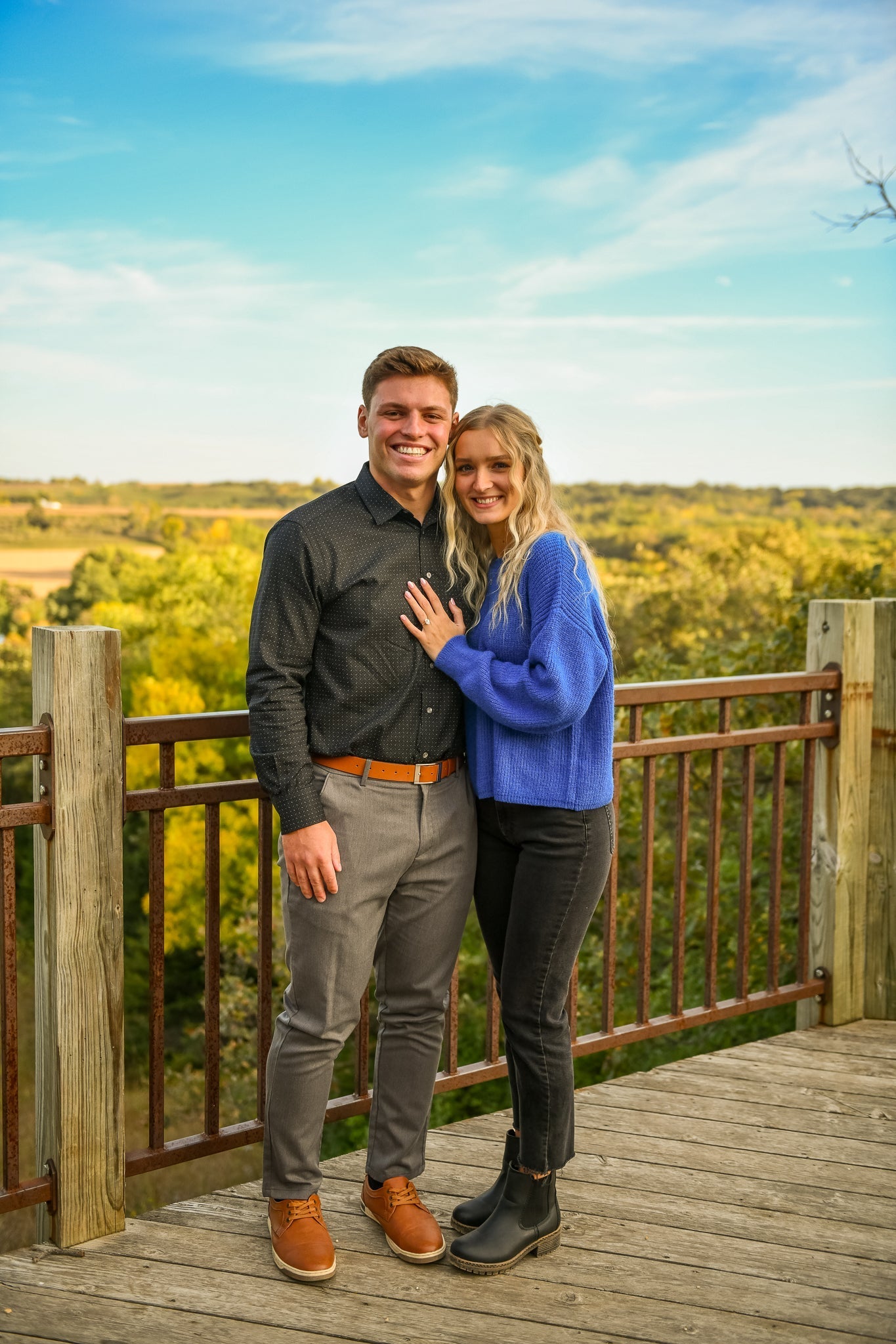 Mallory and Brayden smiling on a wooden overlook, Mallory showing her engagement ring