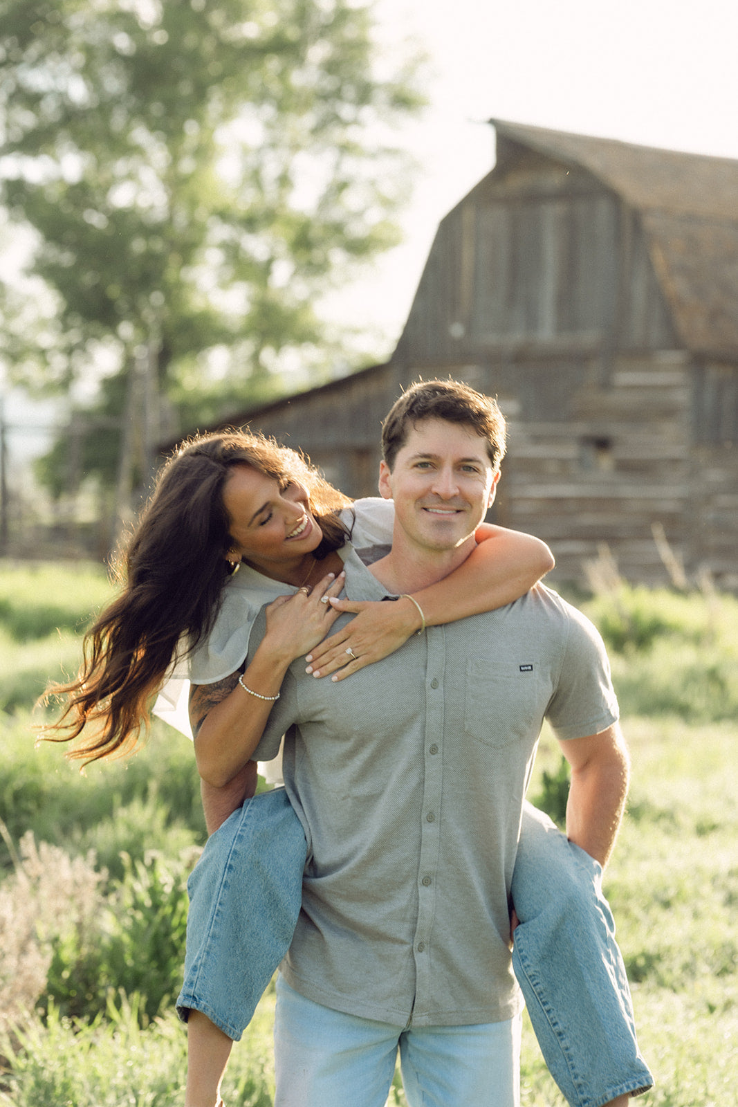 Couple during piggyback in sunlit meadow with rustic barn in background, woman smiling with engagement ring visible