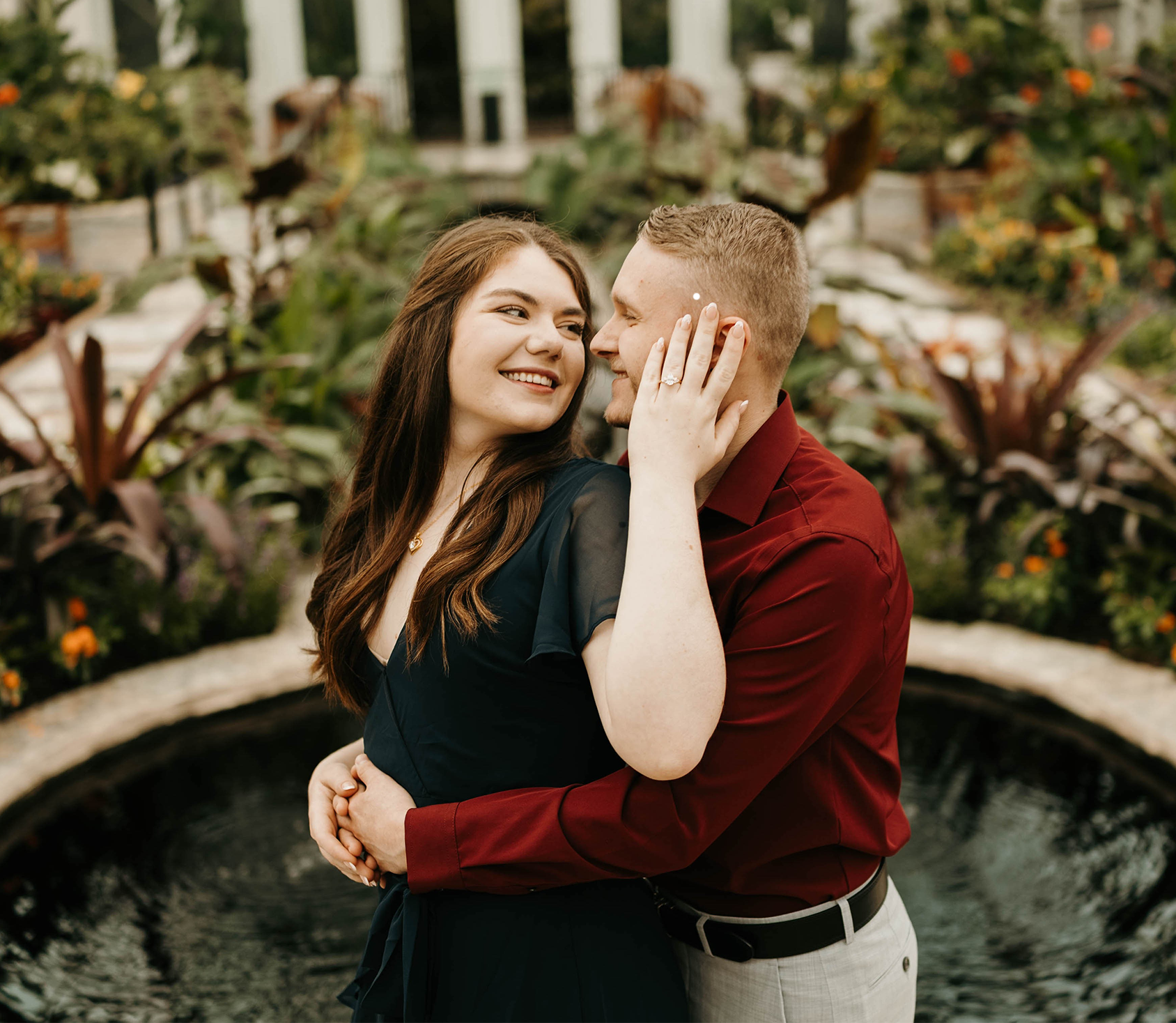 Megan and Kaleb embracing beside a garden fountain, Megan showing her engagement ring and smiling at Kaleb.