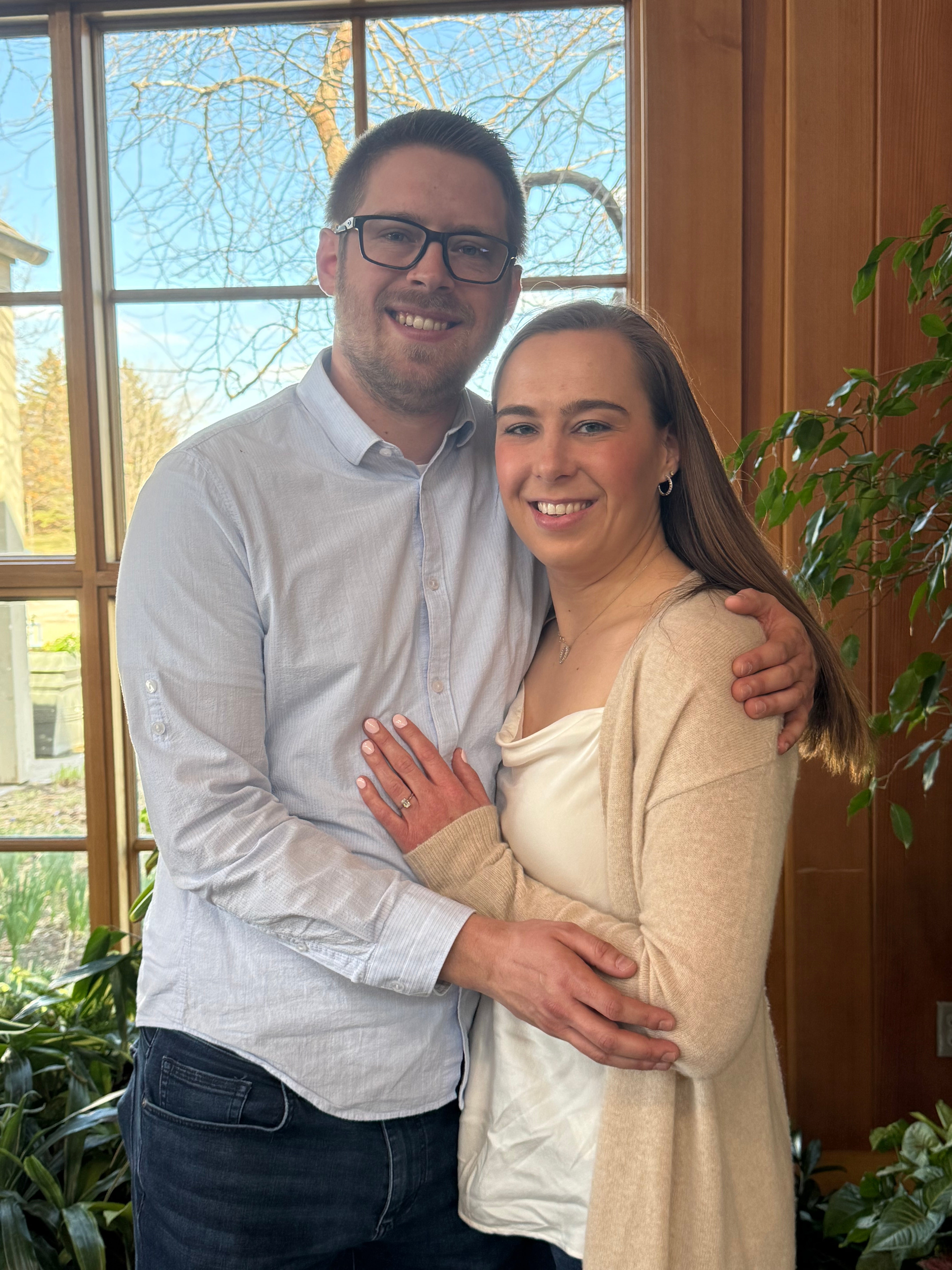 Kelly and Andrew embracing by a window; Kelly shows engagement ring on her left hand.