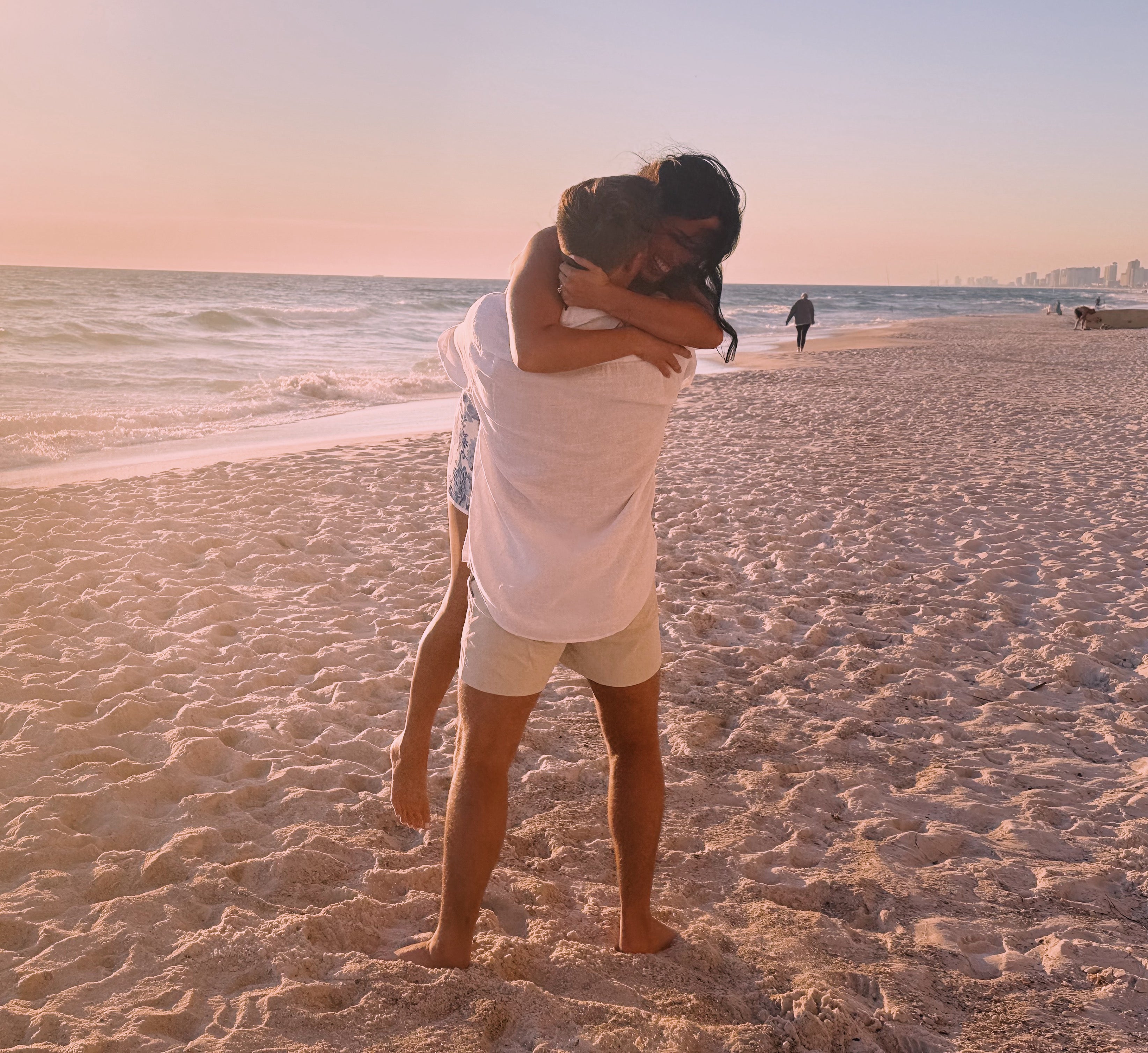 Nevaeh and Colin embracing on a sandy beach at sunset; he lifts her near the shoreline