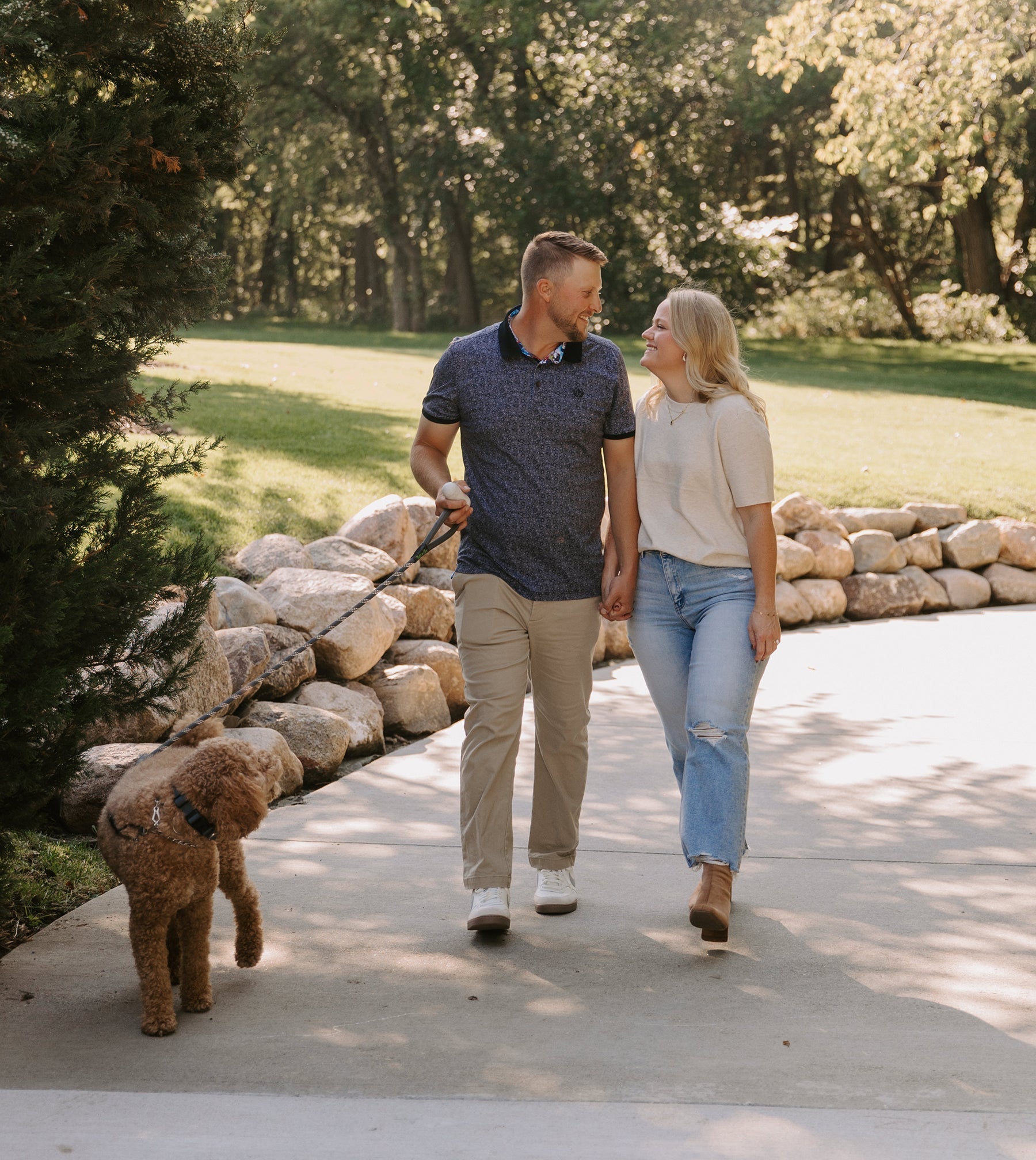Man in a navy polo and woman in a cream top walking hand-in-hand with a brown curly dog along a sunlit park path
