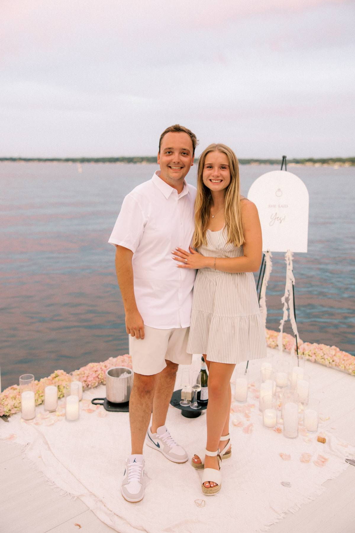 Couple smiling on a lakeside dock at sunset, woman showing her engagement ring beside a sign that reads ‘She said yes!’