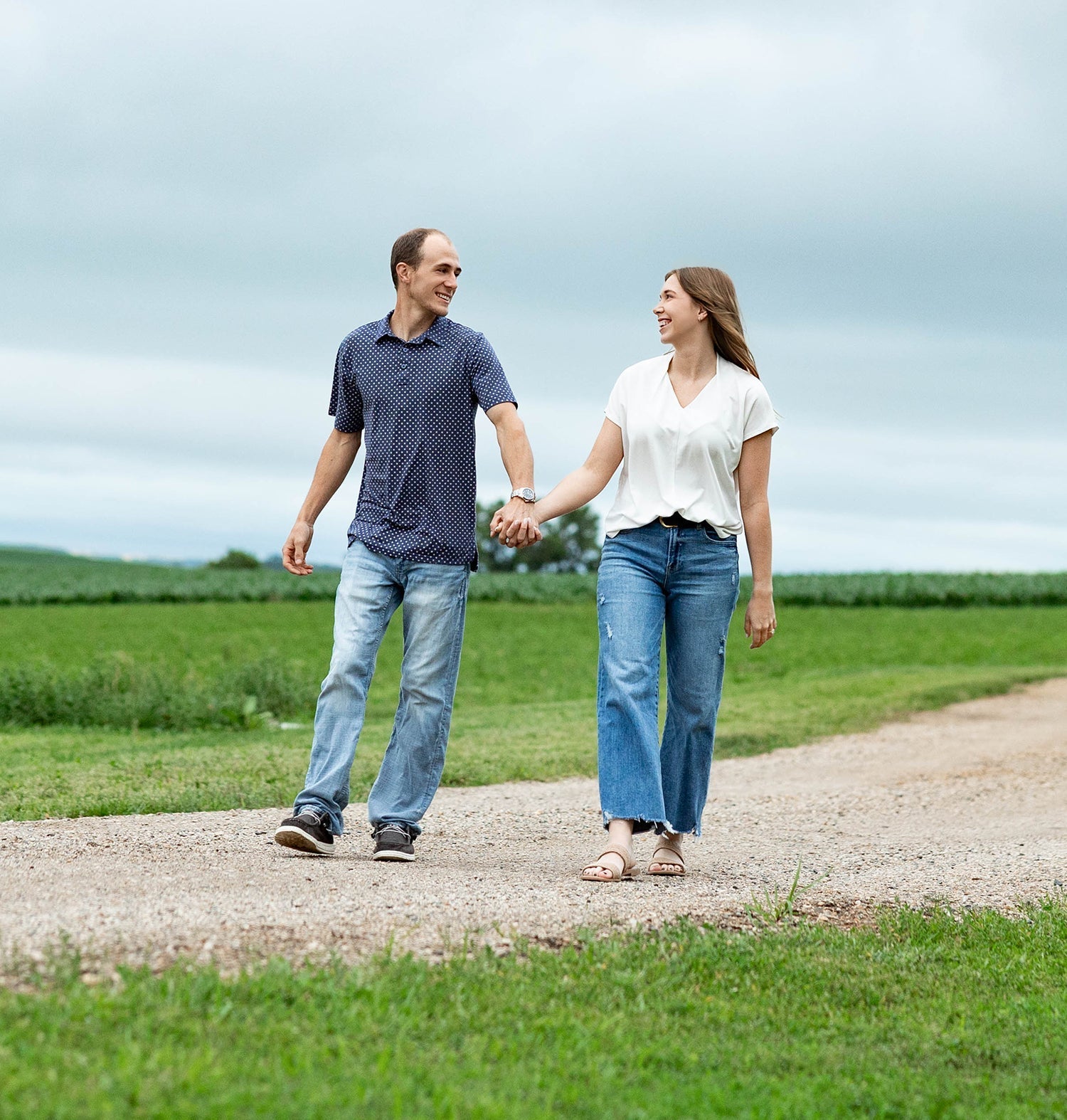 Couple holding hands and smiling as they walk on a gravel road past green fields under a cloudy sky