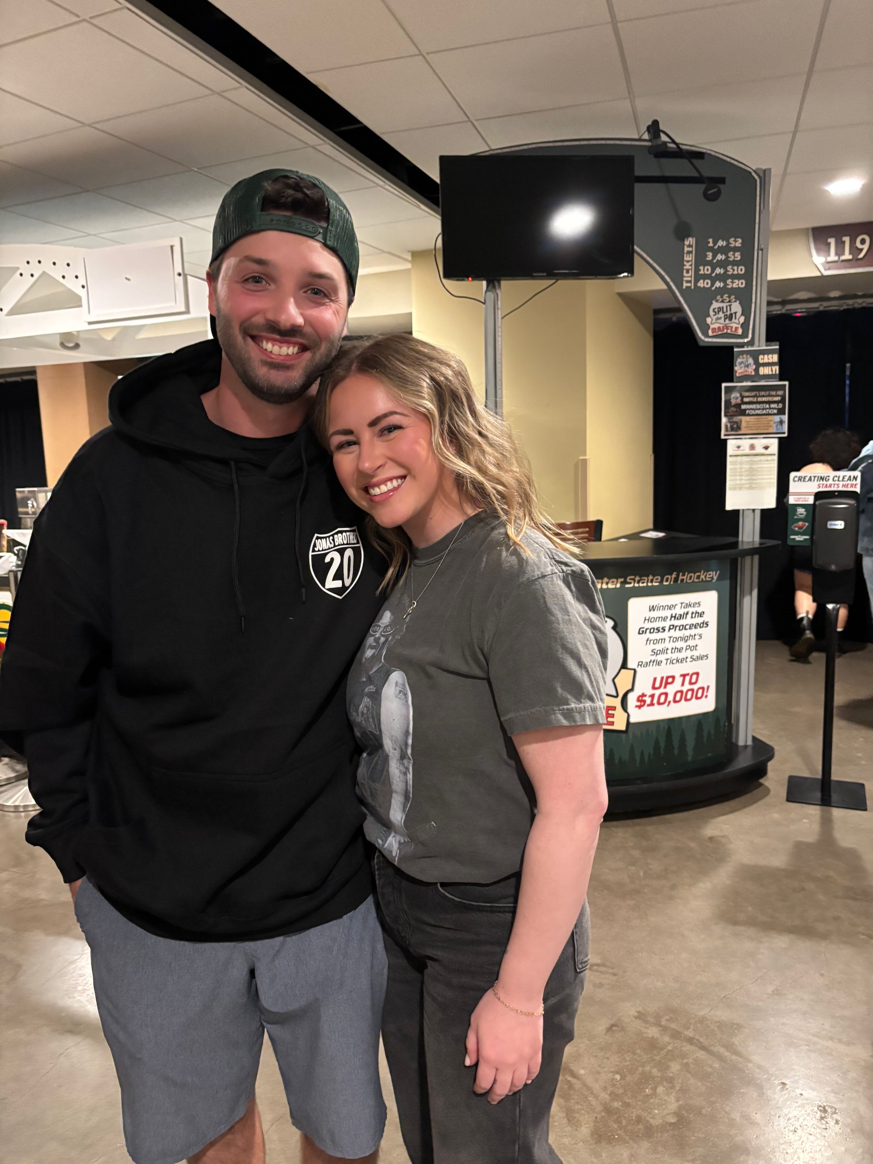 Smiling couple embracing in a concert venue concourse with concession stand and overhead monitor behind them