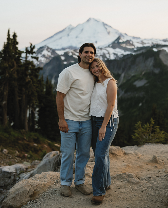 Couple smiling and embracing on a rocky trail at sunset with snow-capped Mount Baker behind them, woman wearing an engagement ring