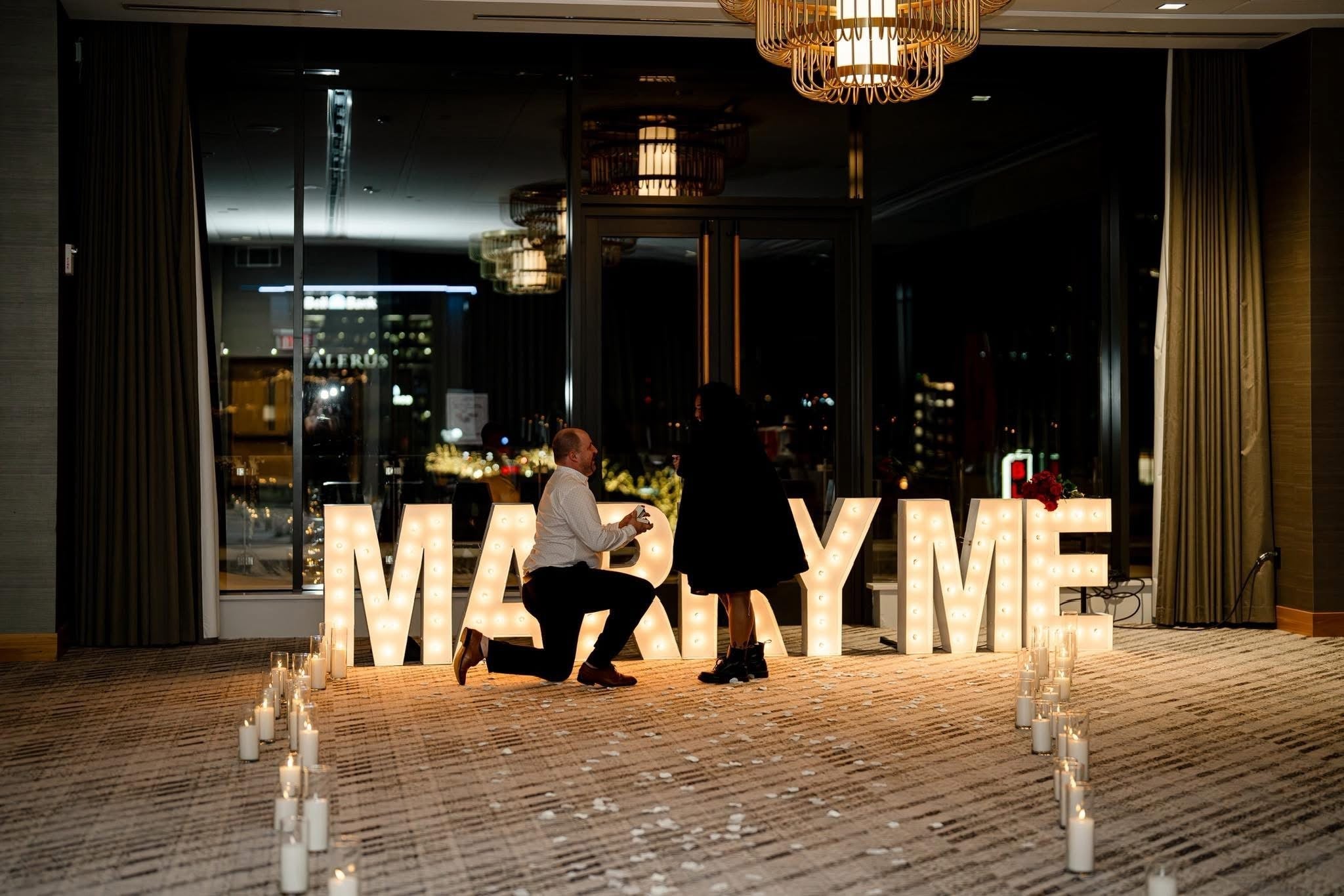 Man kneeling proposing to woman before illuminated MARRY ME sign and candles, city window with ALERUS sign — Alina and Alex