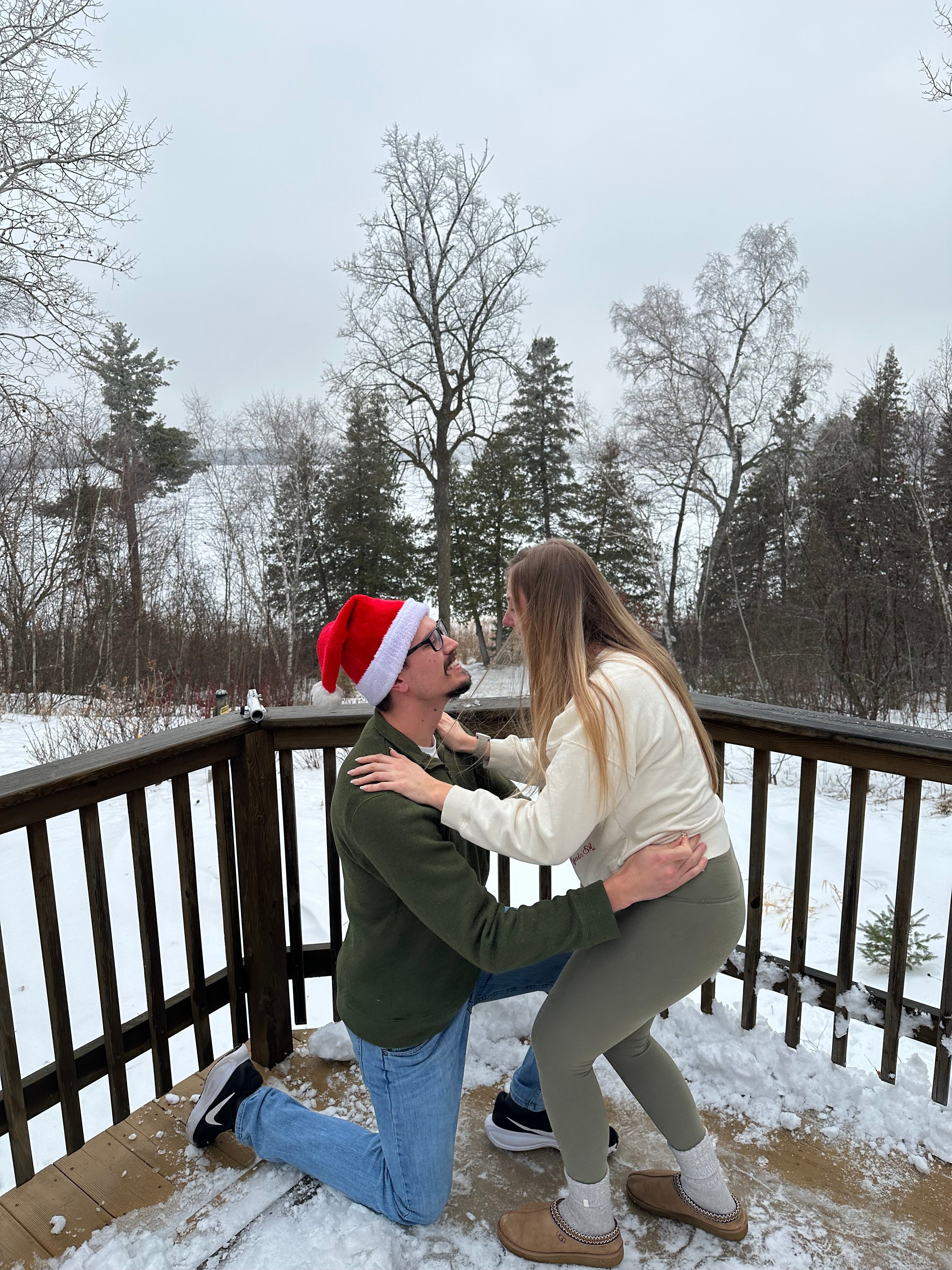 Man in Santa hat kneels to propose to a woman on a snowy deck with bare trees and a frozen lake behind them