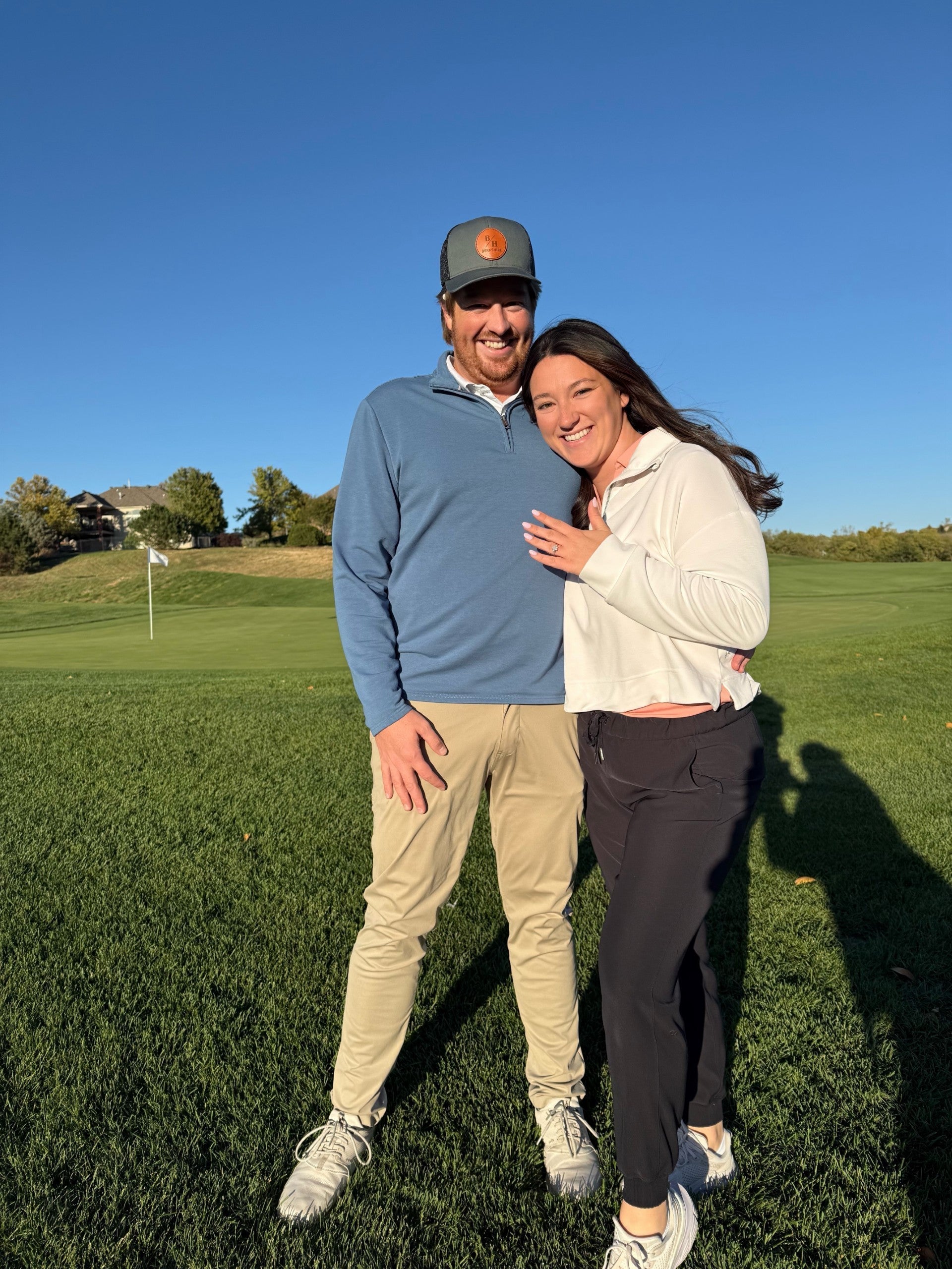 Smiling couple on a golf green after a proposal, woman showing radiant center engagement ring with two round side stones