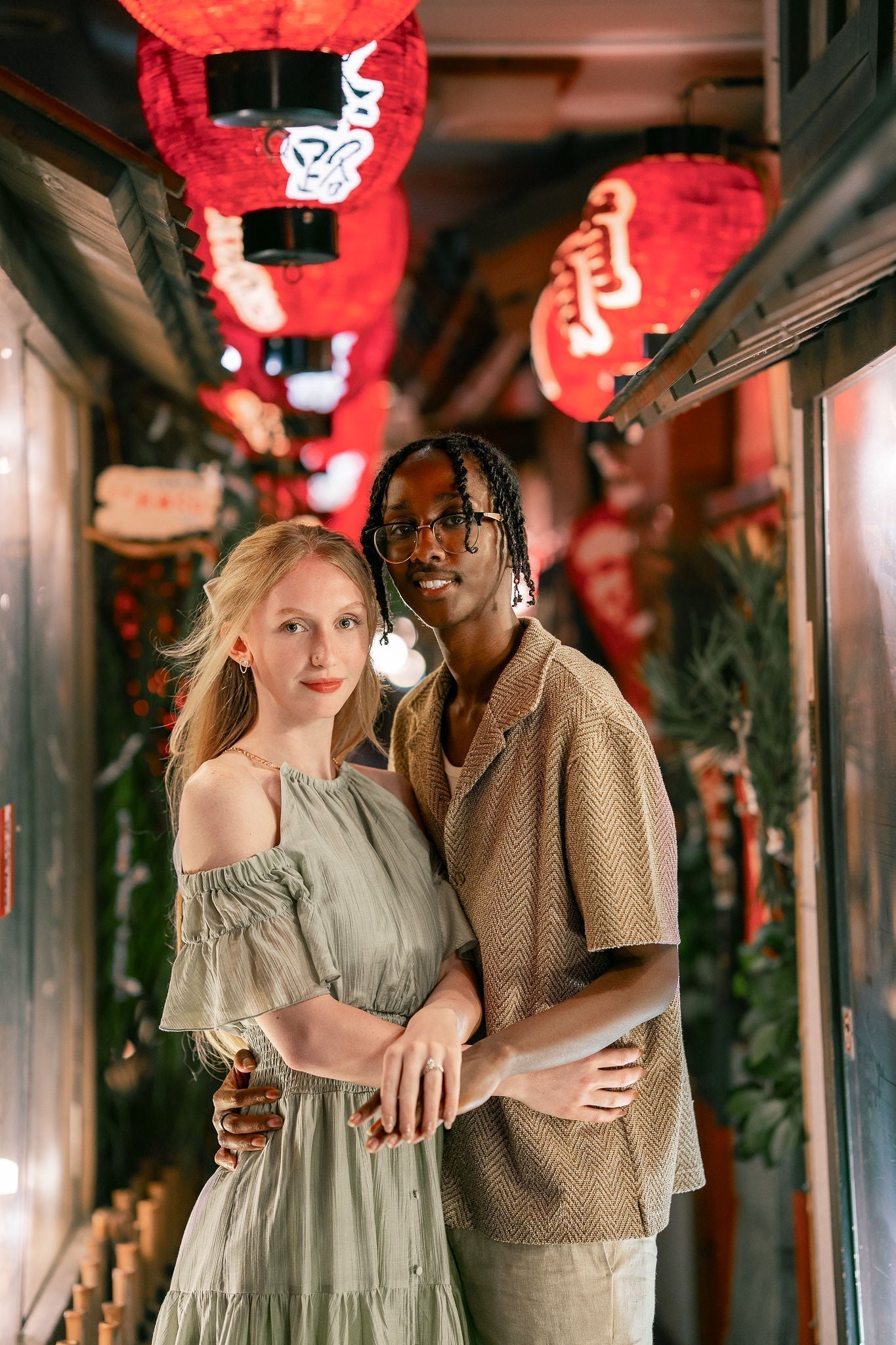 Couple embracing in a narrow lantern-lit alley, woman in a sage green off-shoulder dress and man in a beige short-sleeve shirt