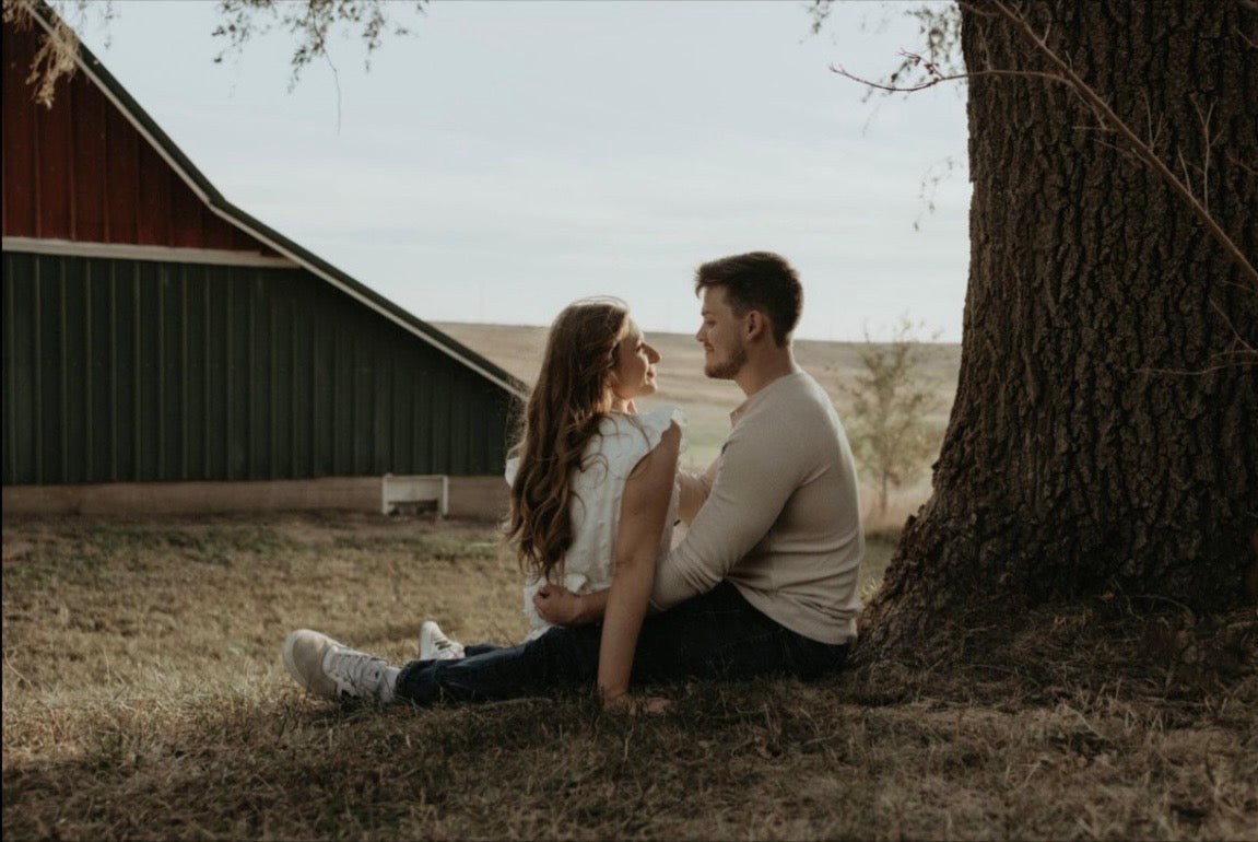 Couple sitting on grass under a tree, embracing and facing each other with a barn and open field in the background
